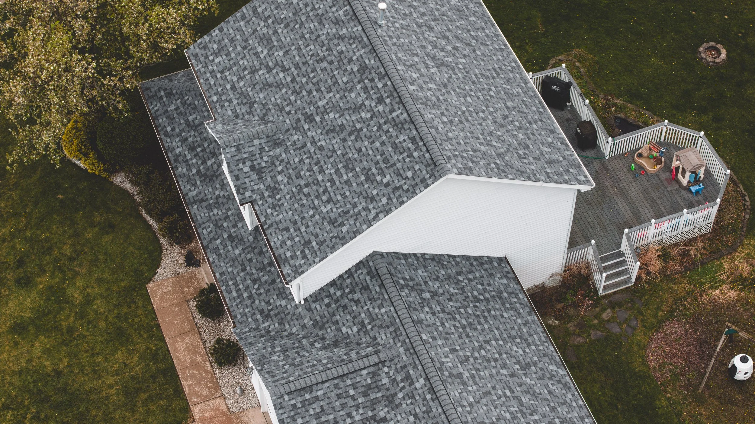 Aerial view of a house with gray shingle roof and a fenced backyard with outdoor play equipment, greenery, and a fire pit.