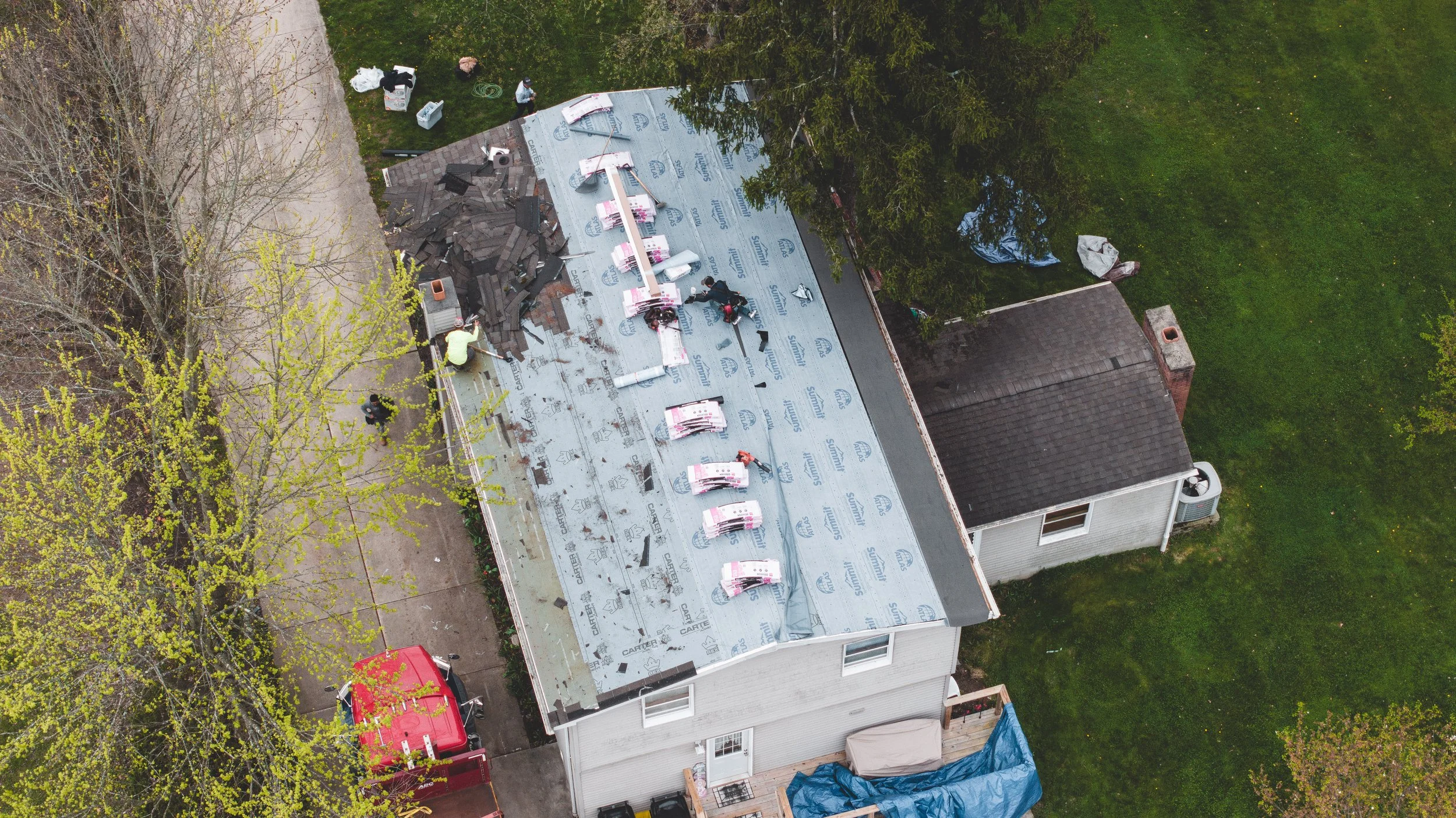An aerial view of a house under roof construction with roofing materials and workers on the roof, surrounded by green trees and a lawn.