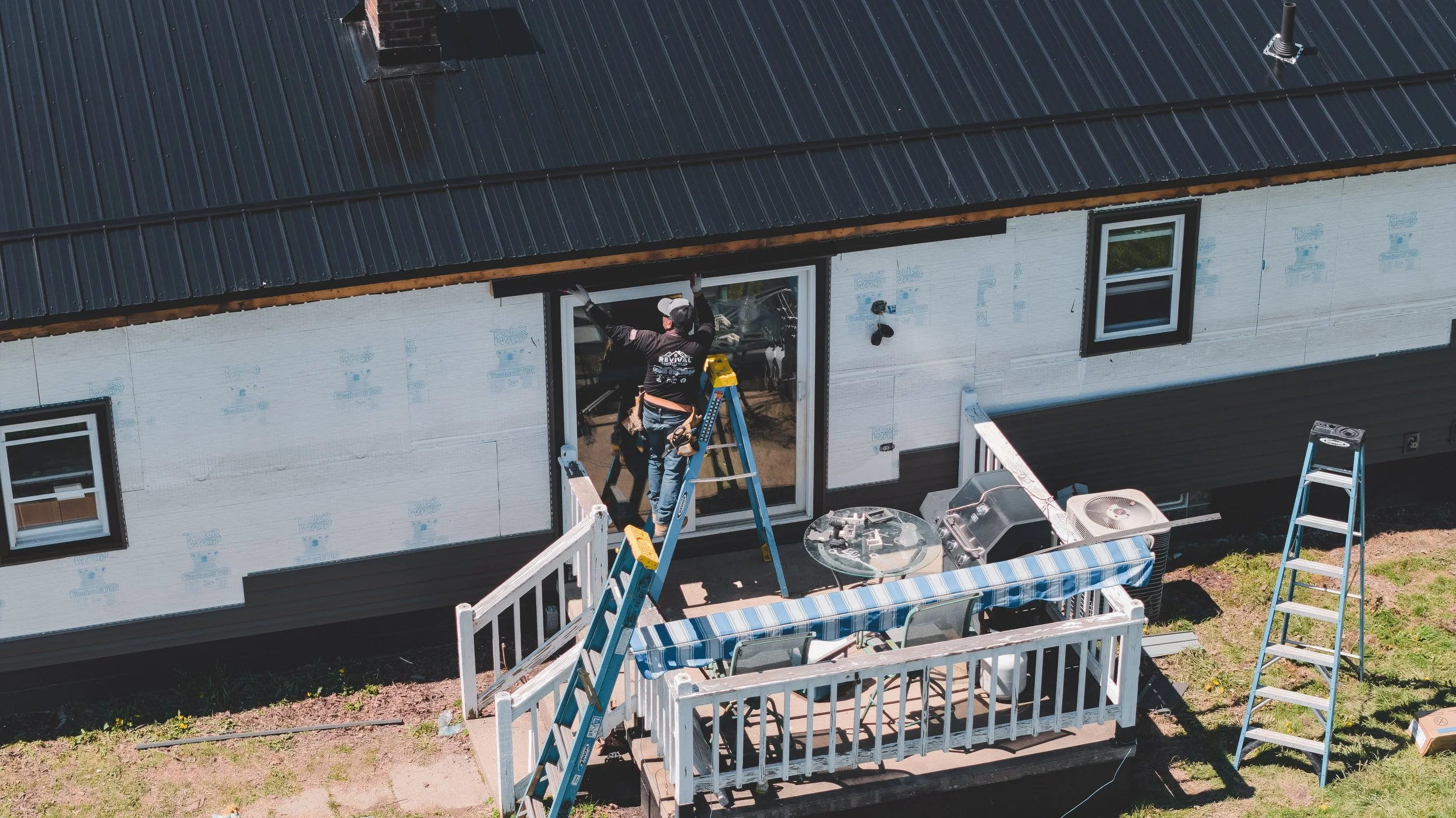 Construction workers installing siding on a house exterior with a black metal roof, two ladders, and outdoor furniture.