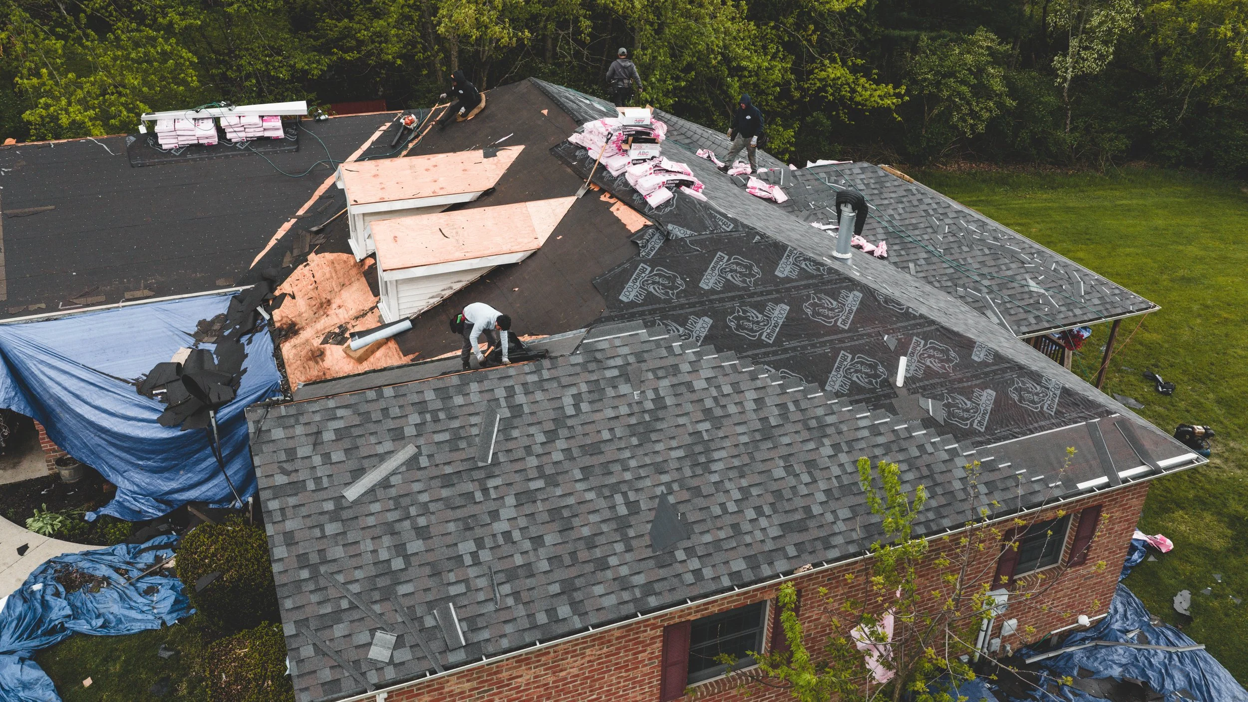 Aerial view of a house roof under renovation with roofing workers installing new shingles, surrounded by green trees and lawn.