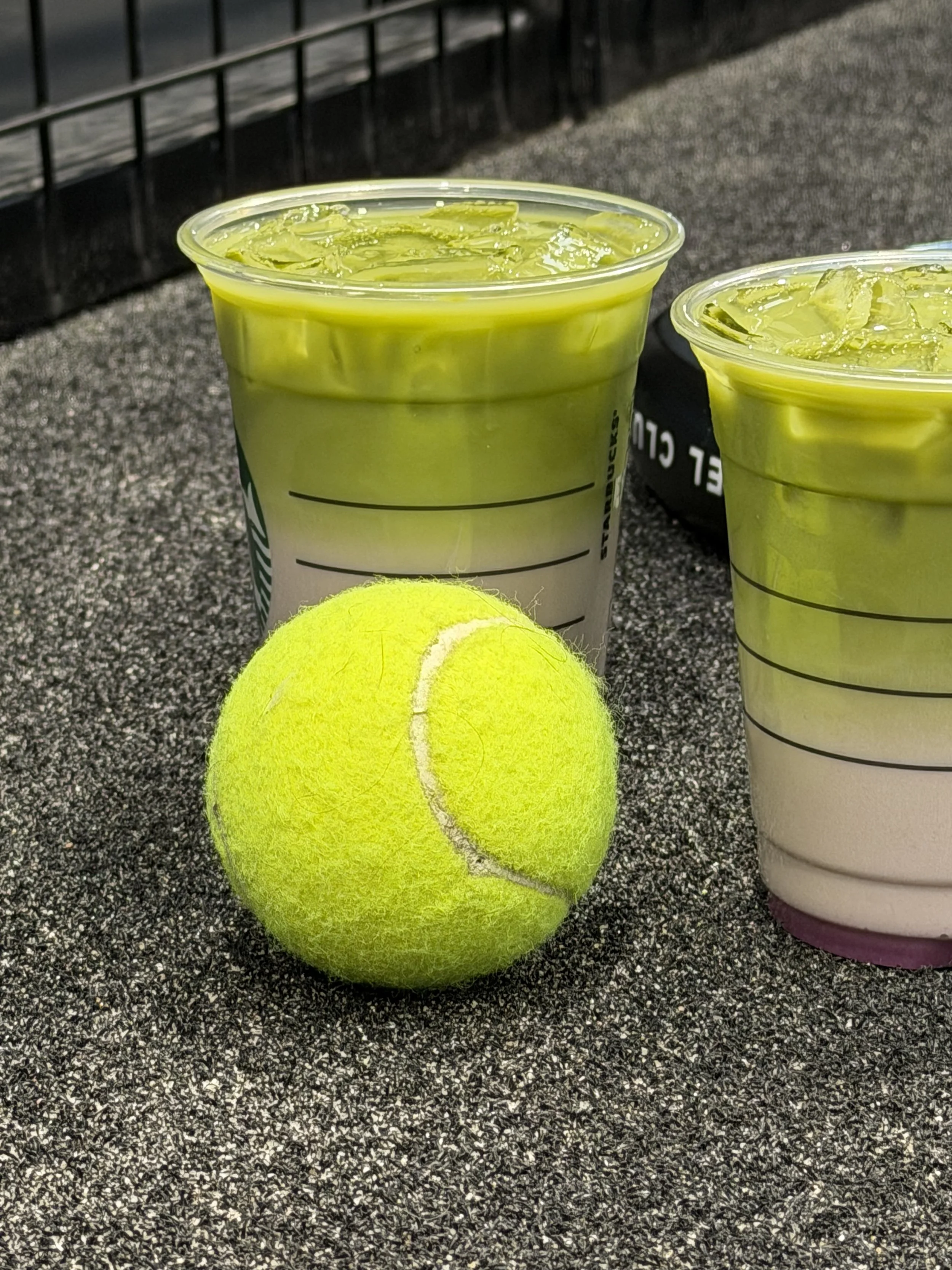 Two Starbucks iced drinks with green tea and a tennis ball resting on a textured surface.