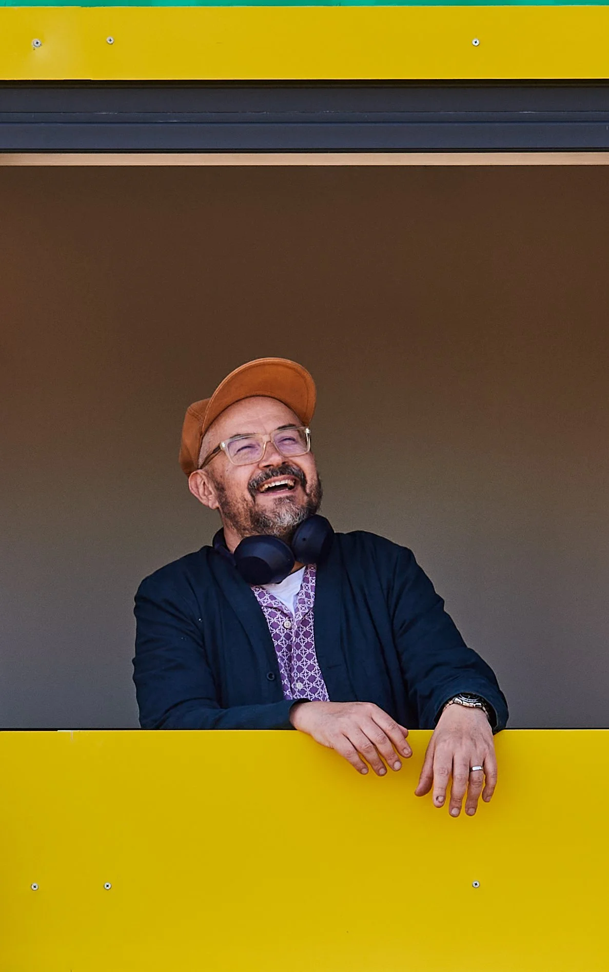 Descriptive: A portrait of Loose FM founder Mark Freeman, wearing an orange cap, glasses, and a dark jacket with headphones around his neck, looking up with a wide smile as he leans on the sill of a vibrant yellow window.