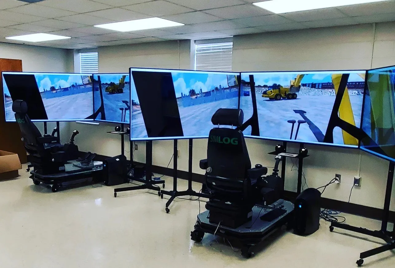 Three large monitors displaying a virtual construction site with a yellow dump truck and construction equipment, situated in an office or training room with simulation chairs and control panels.