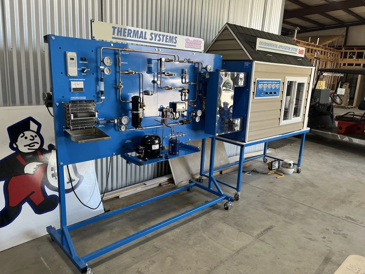 Educational training setup with blue and beige system panels, pipes, gauges, and controls in an industrial workshop setting.