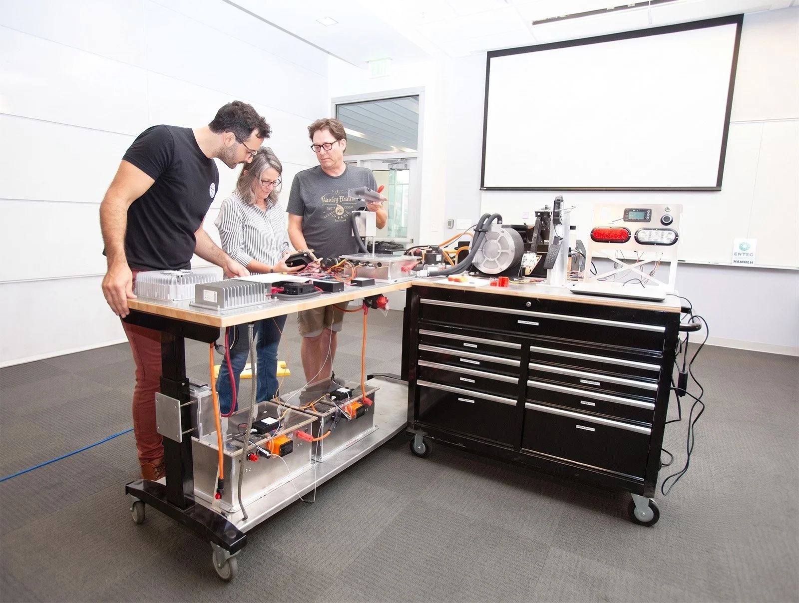 Three people working on a scientific or engineering project in a modern lab with various electronic equipment and a large blank screen on the wall.
