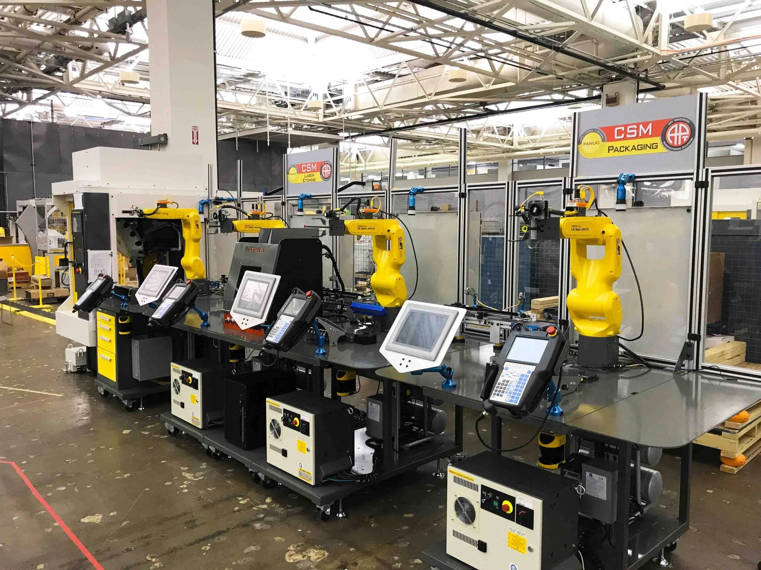 Industrial robotic arms on a workbench inside a manufacturing facility, with control panels and computer monitors in front. Signage for CSM Packaging and laser etching visible in the background.