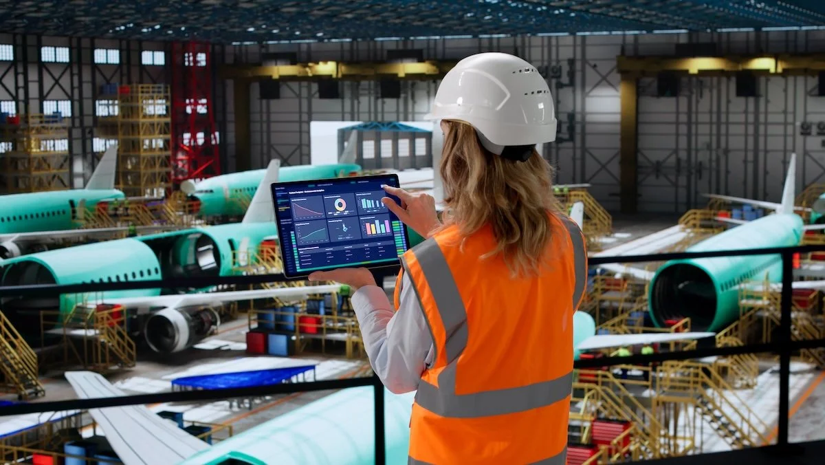 A woman wearing an orange safety vest and a white hard hat monitors data on a tablet inside an airplane assembly hangar with partially assembled airplanes and scaffolding.