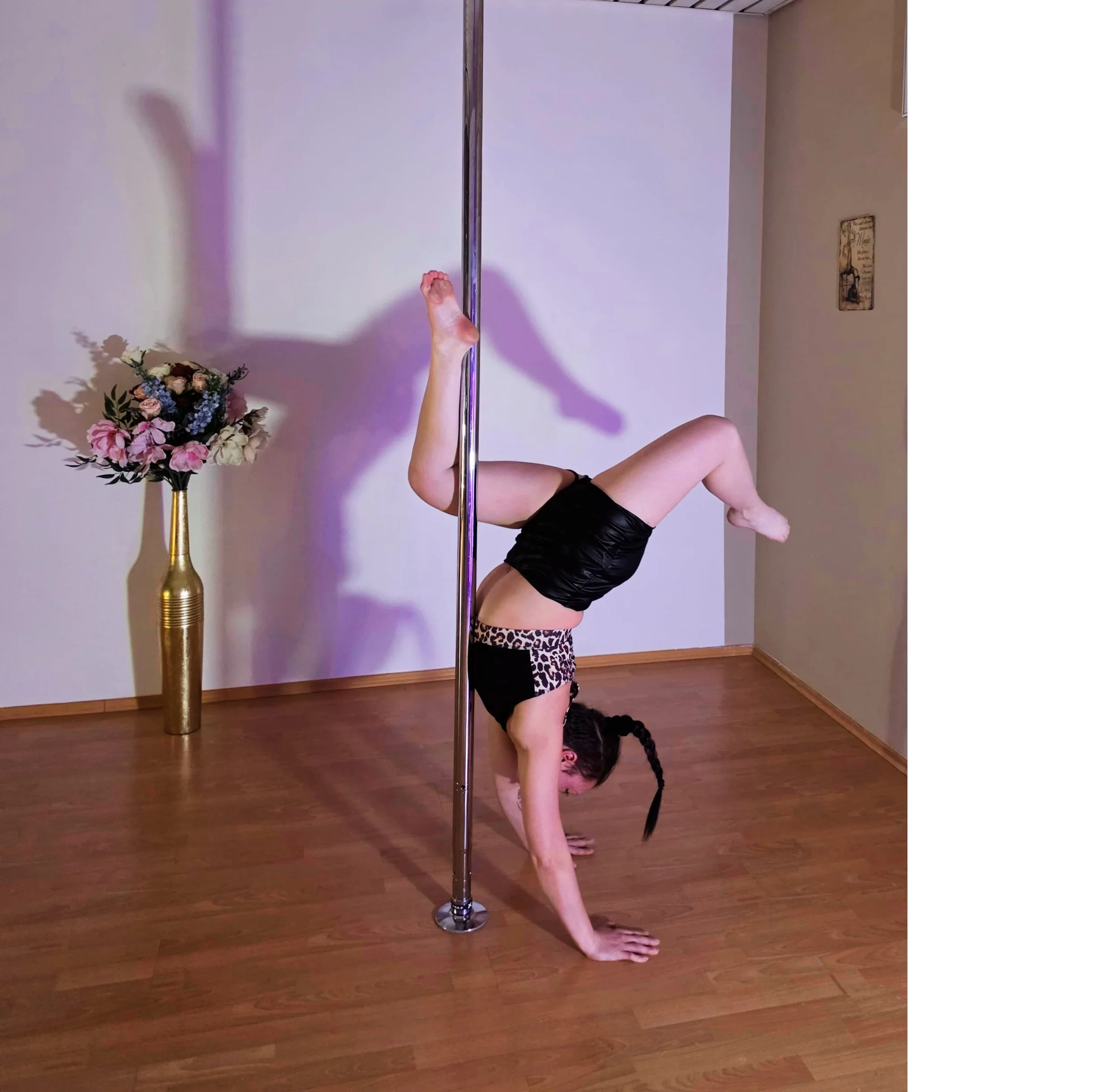 Eine Frau führt eine Pole-Dance-Übung aus, steht auf den Händen mit den Füßen an der Stange, in einem Raum mit Holzboden und einer Vase mit Blumen im Hintergrund.