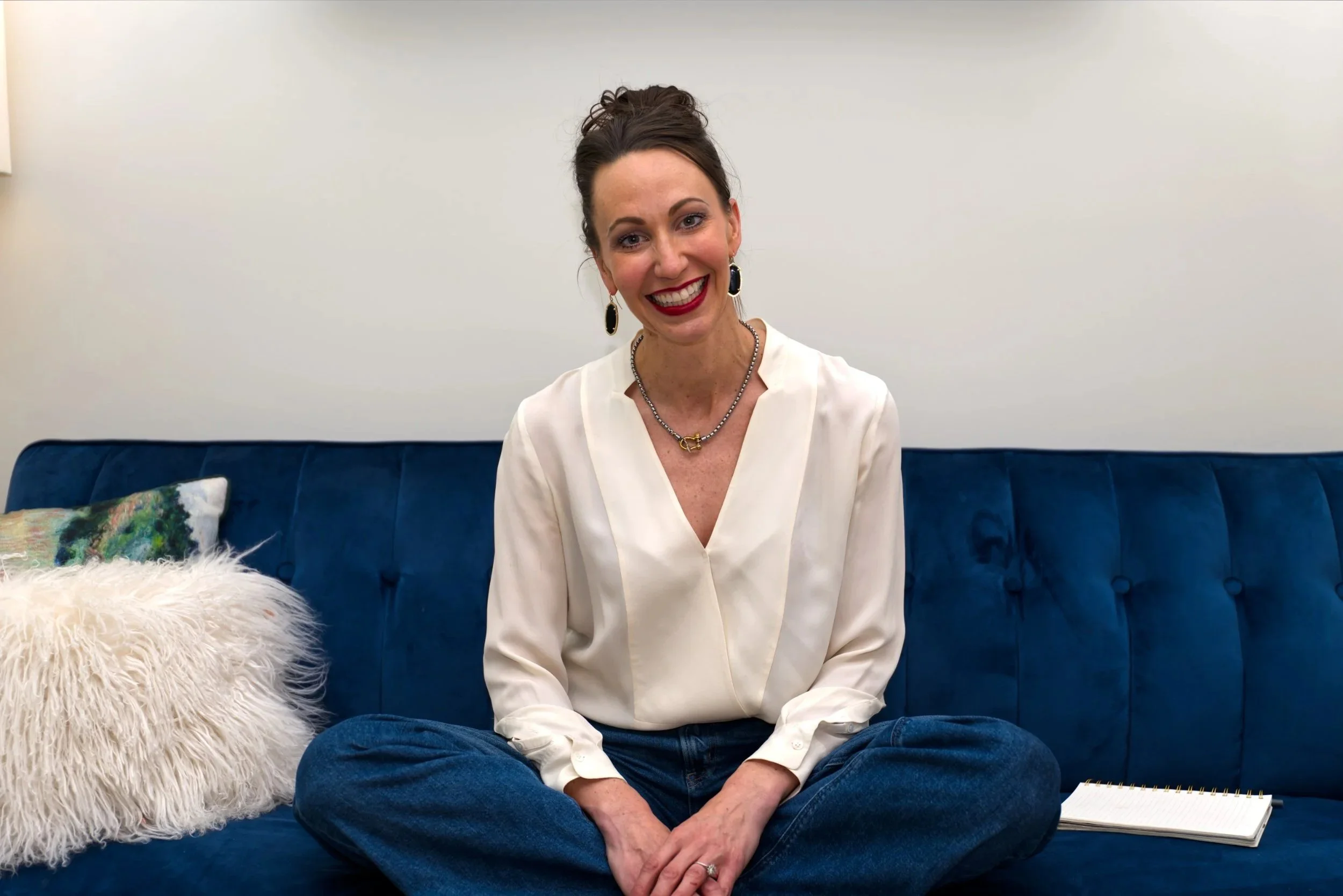 A woman with dark hair in an updo, wearing a white blouse and jeans, sitting cross-legged on a blue velvet couch. She is smiling brightly, showing her teeth, with earrings, a necklace, and a ring. There is a fluffy white pillow and a colorful pillow on the left side of the couch, and a notebook on the right side.