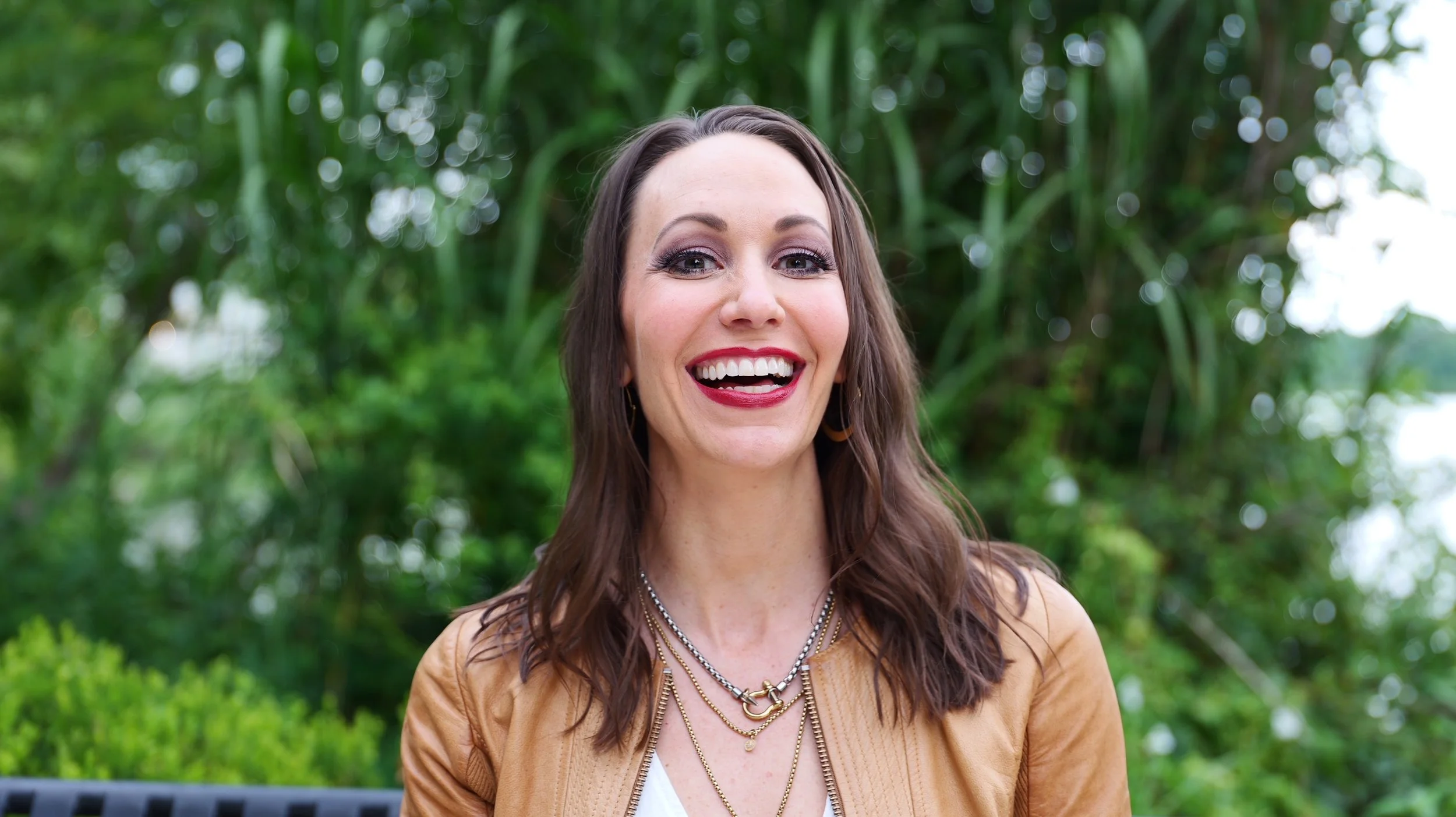 Woman with long brown hair, wearing a tan jacket and layered necklaces, smiling outdoors with greenery in the background.