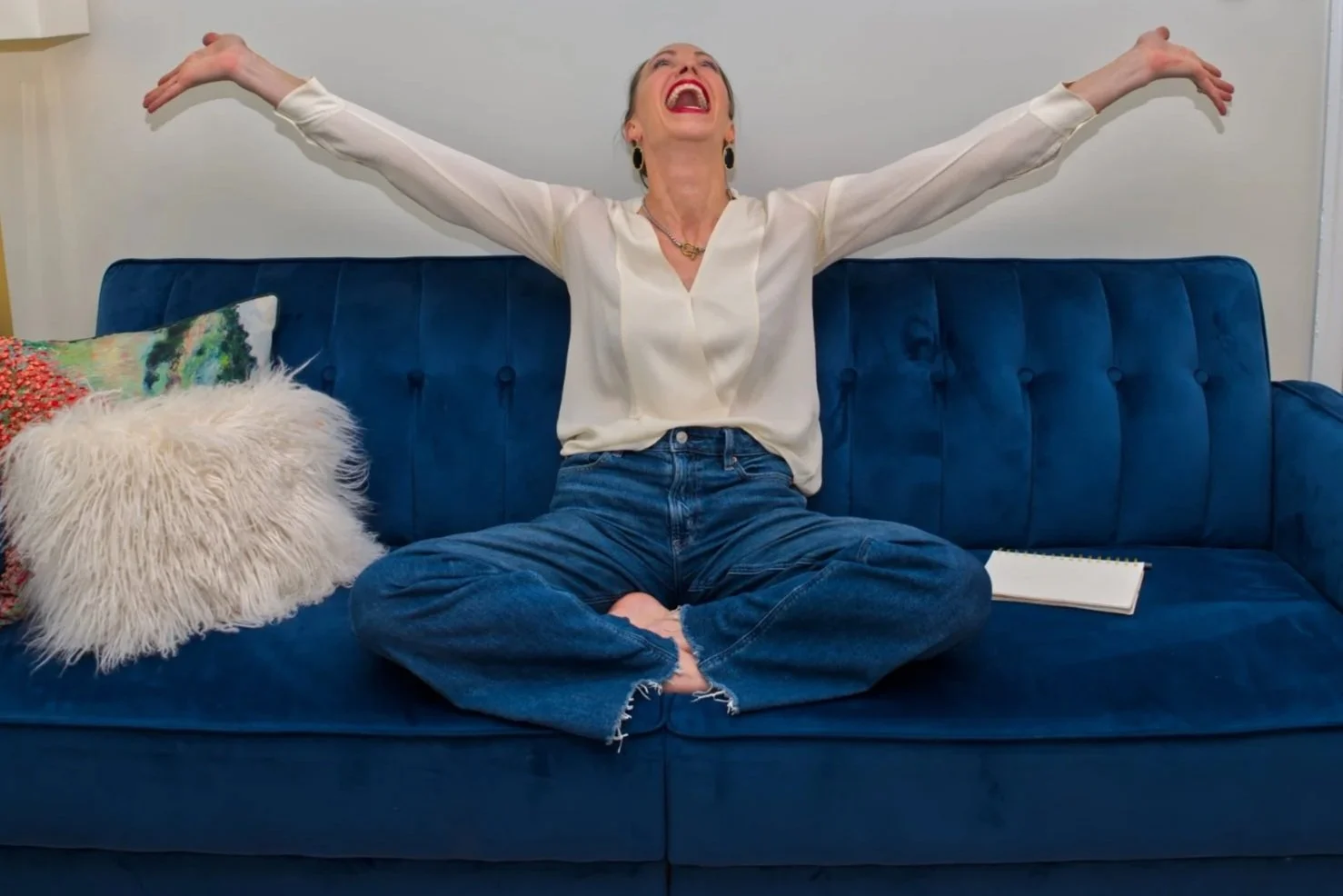A woman sitting cross-legged on a blue velvet sofa, stretching with arms out wide, smiling and laughing, with a notebook resting on the sofa beside her, and decorative pillows behind her.