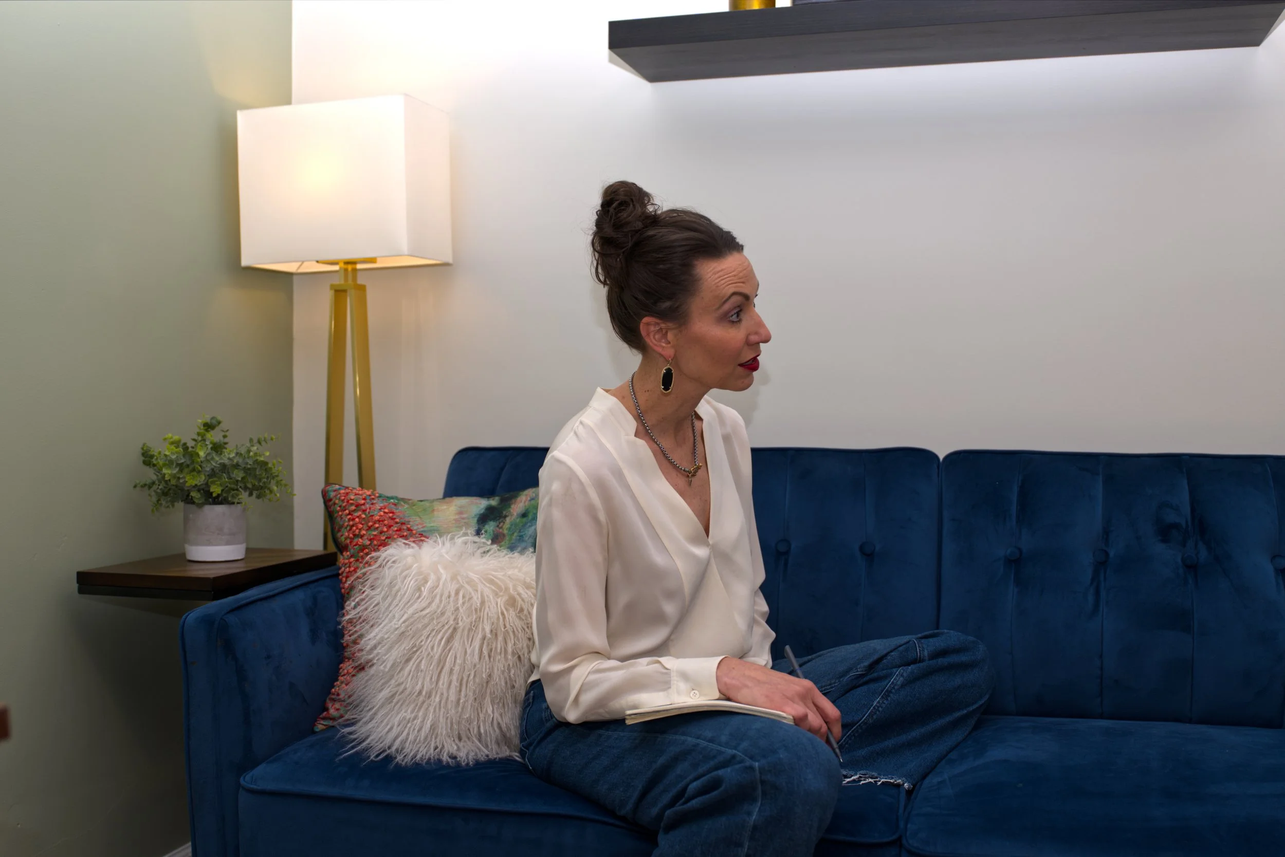 A woman with dark hair in a top bun, wearing a white blouse, earrings, and a necklace, sitting on a dark blue sofa in a living room. She is holding a notepad and pen, with a decorative pillow and a fluffy white throw on the sofa. Behind her, there is a side table with a potted plant, a tall floor lamp, and a wall shelf with objects. The room has light-colored walls and modern decor.