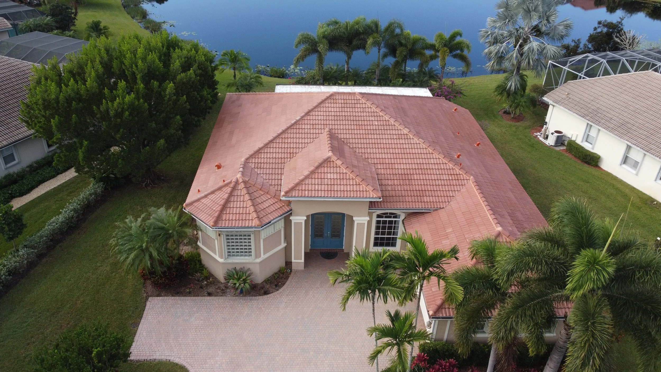 Aerial view of a house with a gray roof, surrounded by tropical trees and neighboring houses with tiled roofs, parked cars in the driveway, and a backyard with a pond or small pool.