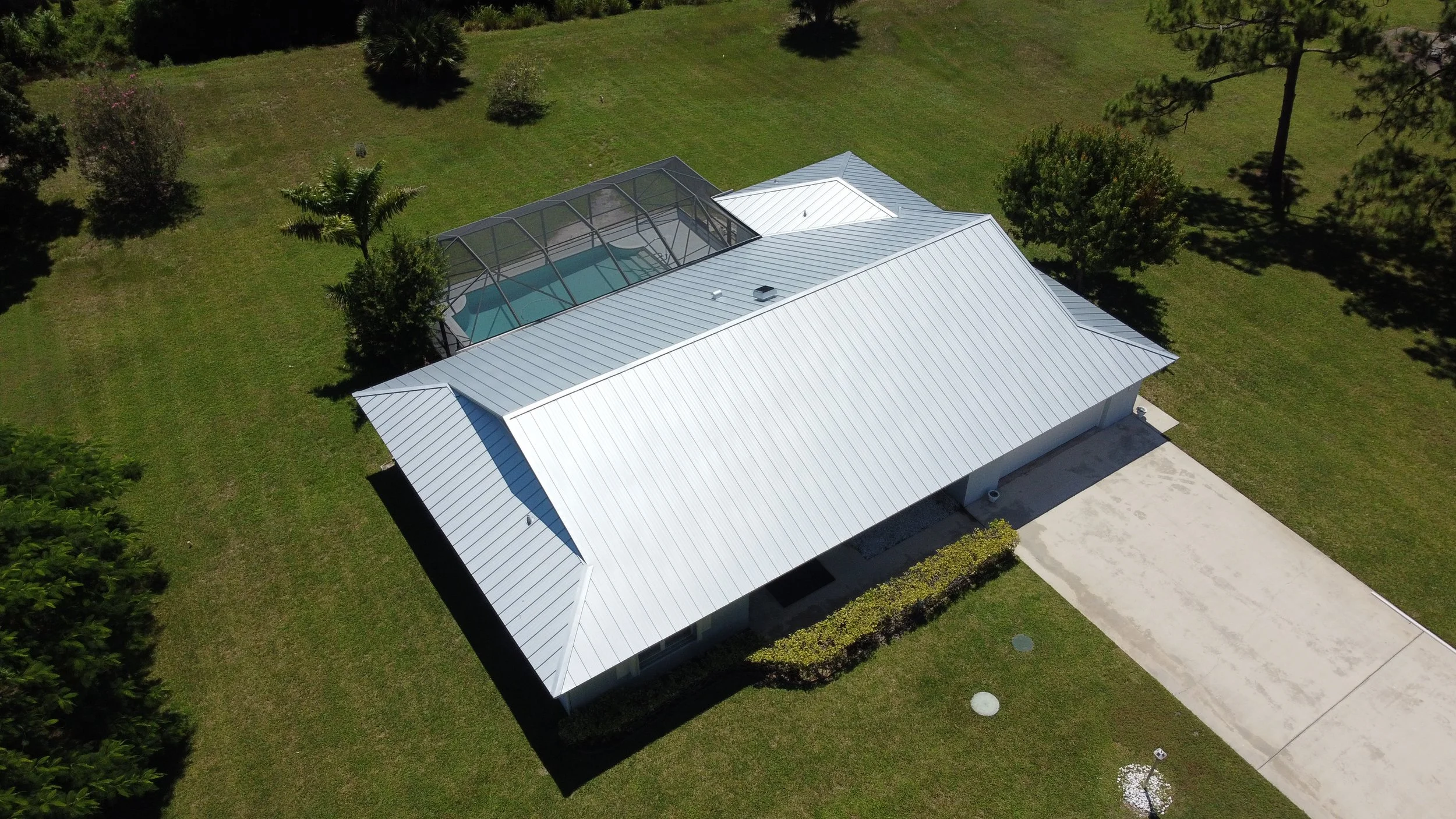 An aerial view of a house with a brown metal roof, a swimming pool, and surrounding palm trees in a backyard.