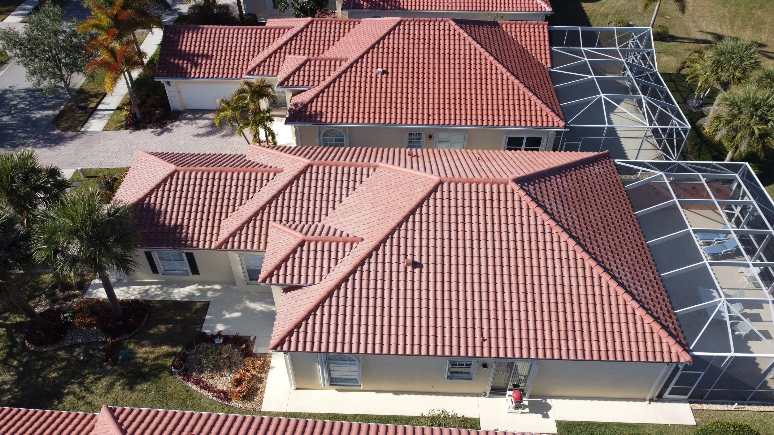 Aerial view of a house with a terracotta tiled roof, a driveway with a red car parked, surrounded by greenery including palm trees, and a screened swimming pool in the backyard.