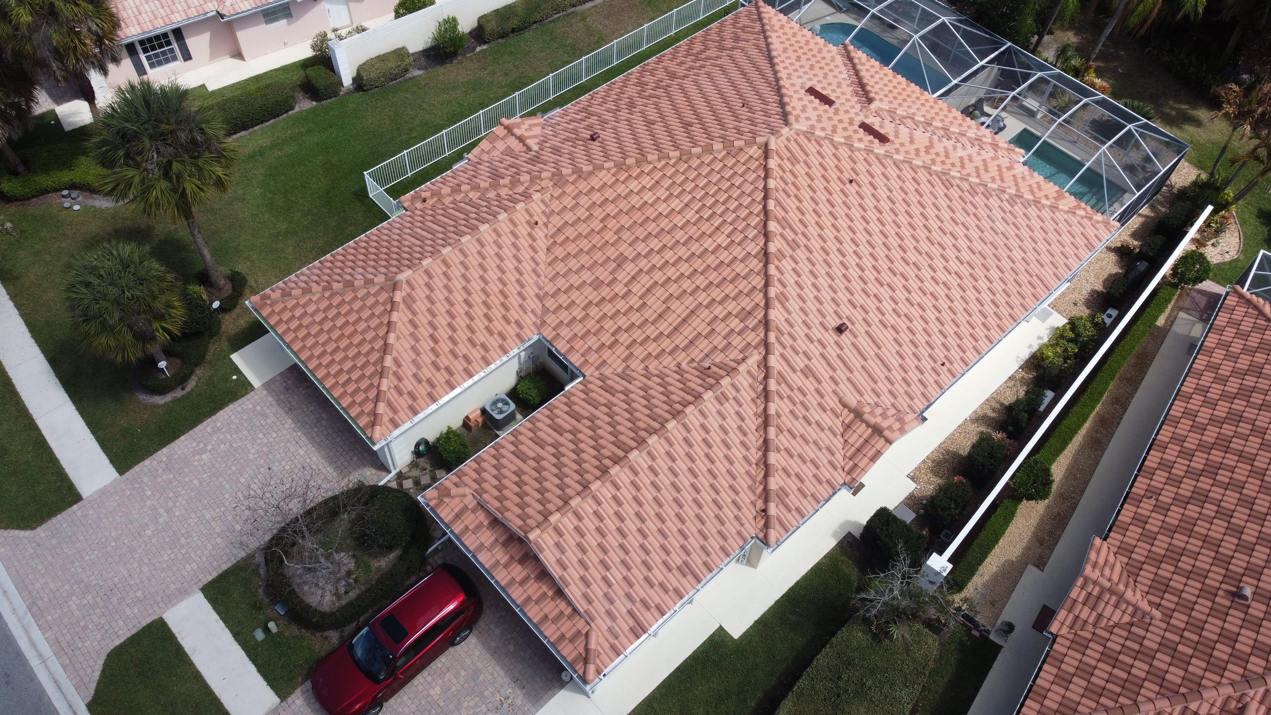 Aerial view of a house with a tan tiled roof, enclosed backyard with a pool, lush green grass, trees, and a driveway with a red car parked on it.