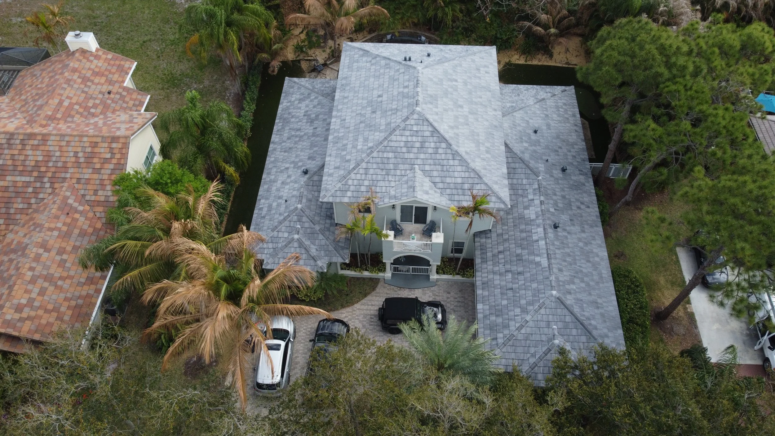 Aerial view of a house with a gray tiled roof, surrounded by palm trees and other greenery, with cars parked in the driveway.