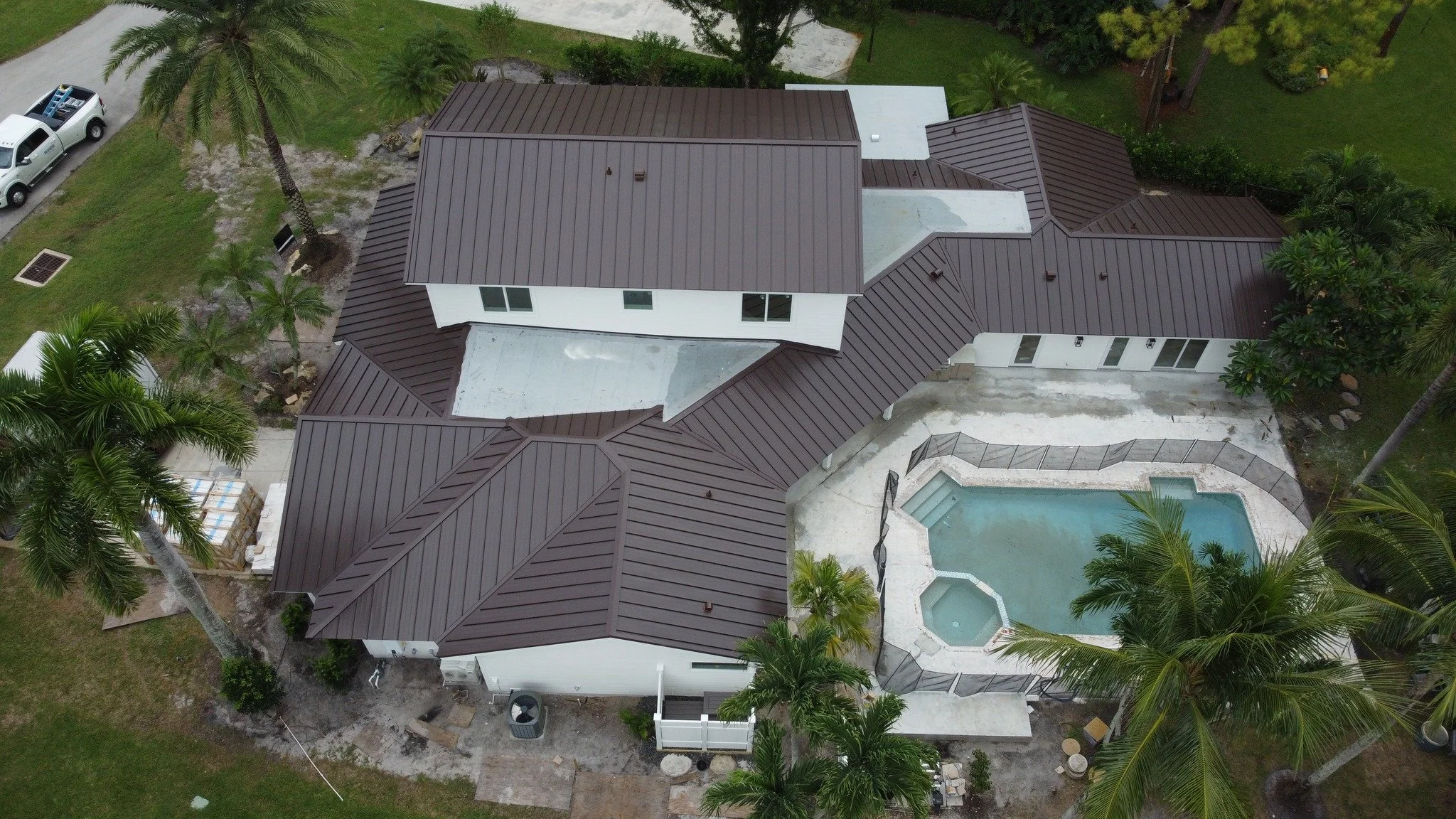 Aerial view of a house with a brown metal roof, surrounded by lush green trees including palm trees, with a backyard swimming pool and patio area.