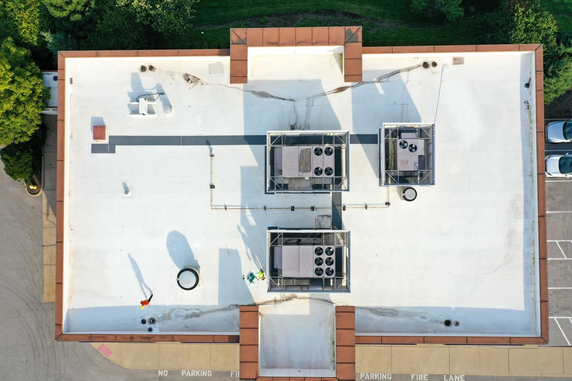 Aerial view of a flat white rooftop with three HVAC units in metal enclosures, vents, and a worker cleaning or inspecting the roof near a black circular object, with a small section of parking lot and trees visible at the edges.