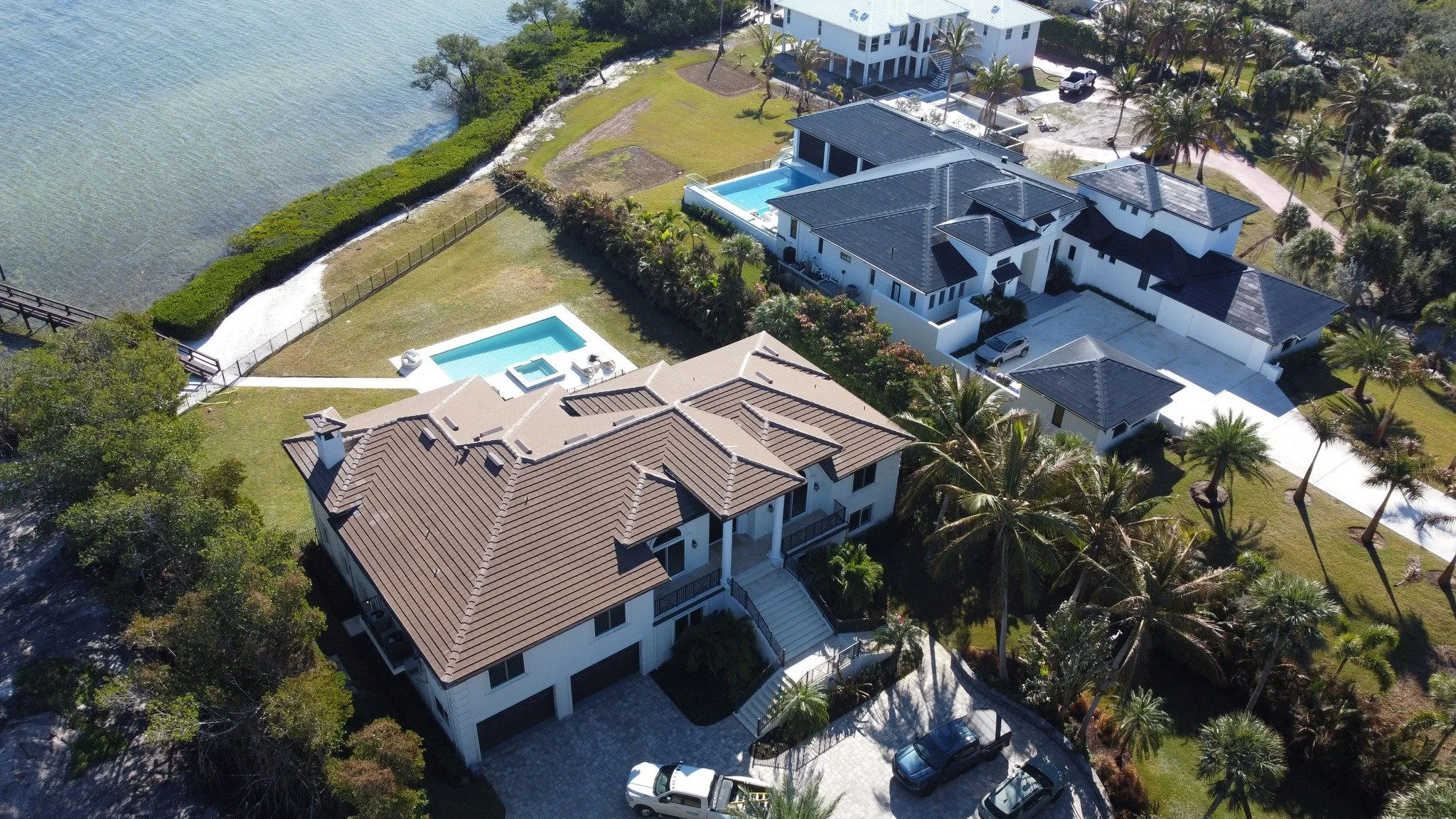 Aerial view of a coastal residential property featuring a large white house with a brown roof, multiple swimming pools, lush tropical landscaping, and water access via a pathway and dock leading to a body of water.