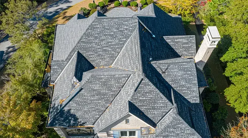 Aerial view of a large house with multiple gray shingle roofs, surrounded by trees and a yard.