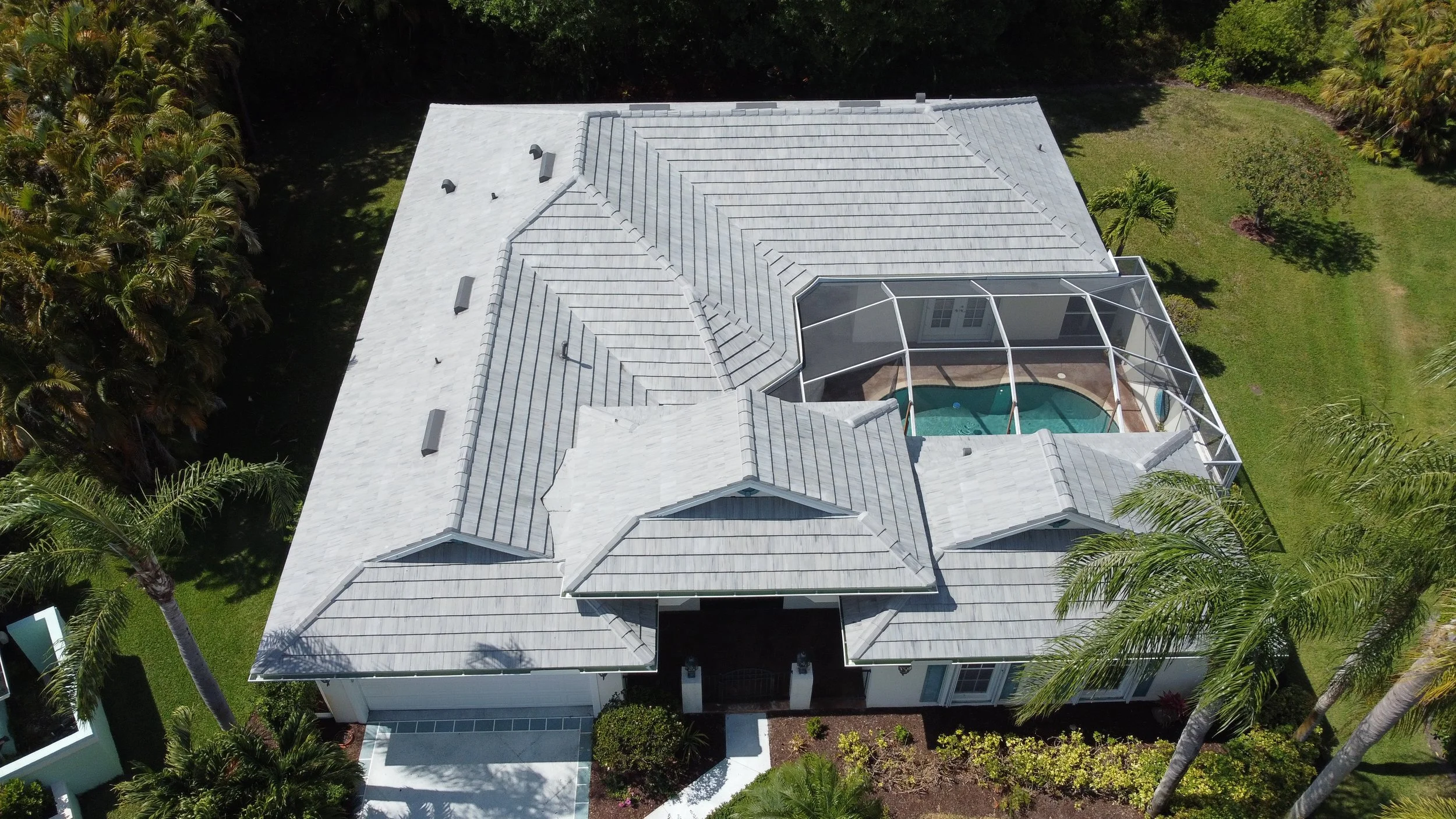 Aerial view of a waterfront residential neighborhood with large houses, swimming pools, lush greenery, palm trees, and a body of water nearby.