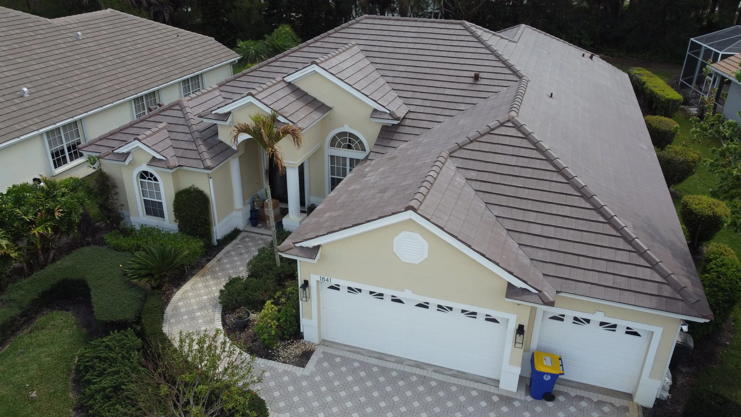 Aerial view of a yellow suburban house with a brown tiled roof, front driveway, and surrounding lush greenery.