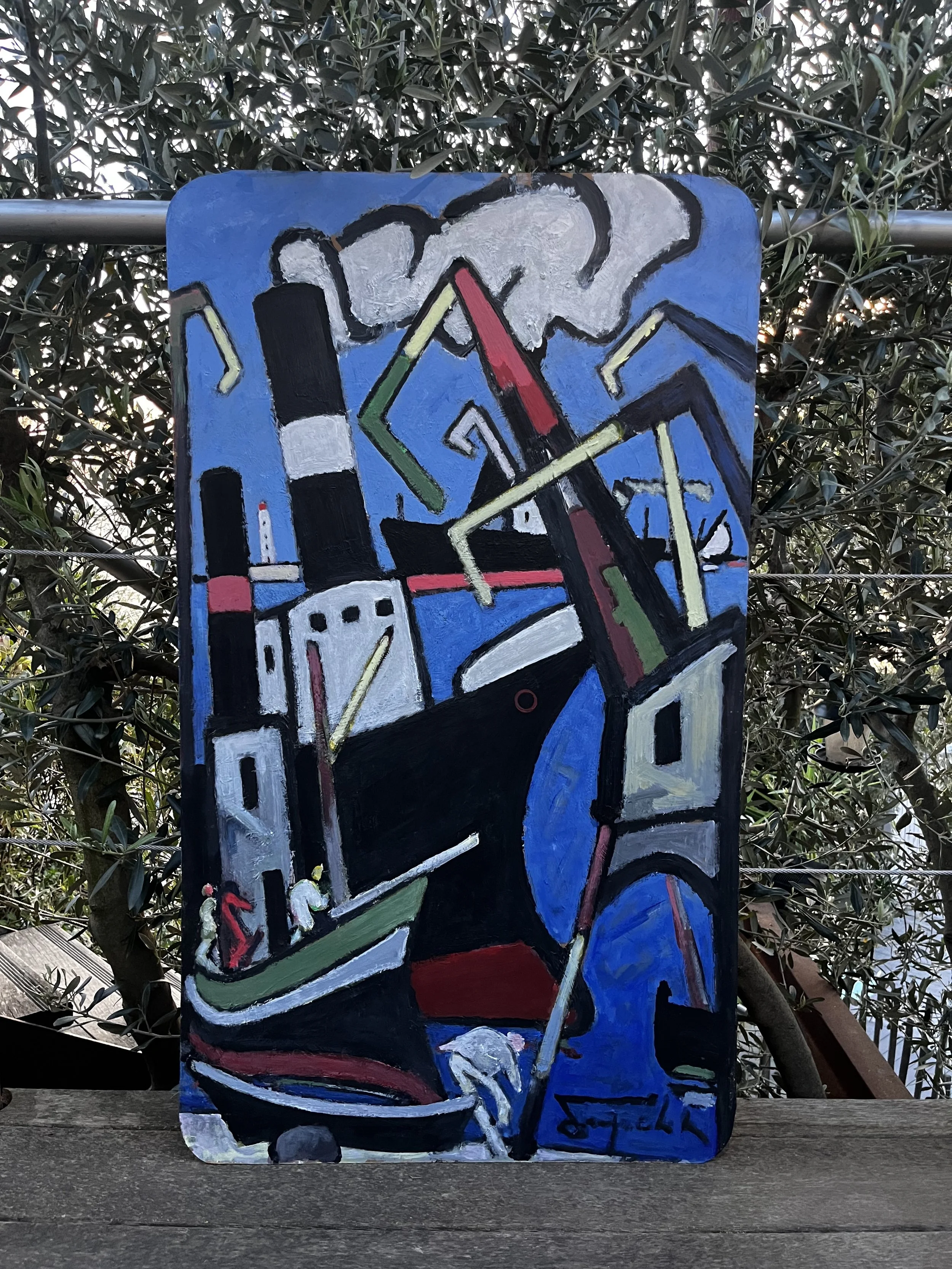 Une peinture abstraite représentant une main tenant un pinceau, avec un fond bleu et des éléments colorés tels que du vert, du rouge, du blanc, du noir. L'œuvre est peinte sur un panneau en bois placé sur une surface en bois, avec des buissons en arr
