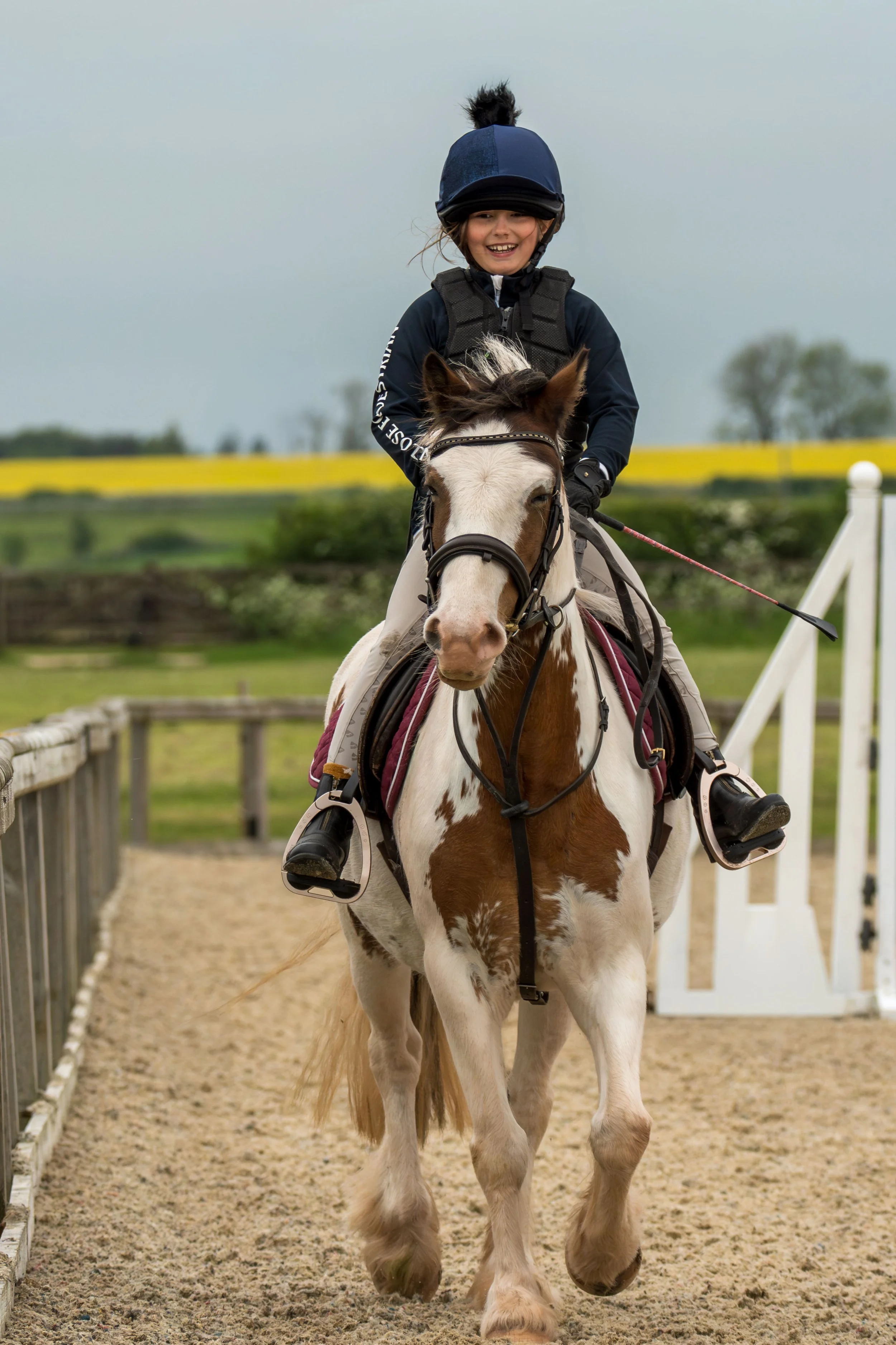 A young girl riding a white and brown horse on a racetrack, wearing a black riding helmet, black gloves, and a black riding jacket, with a cheerful smile.