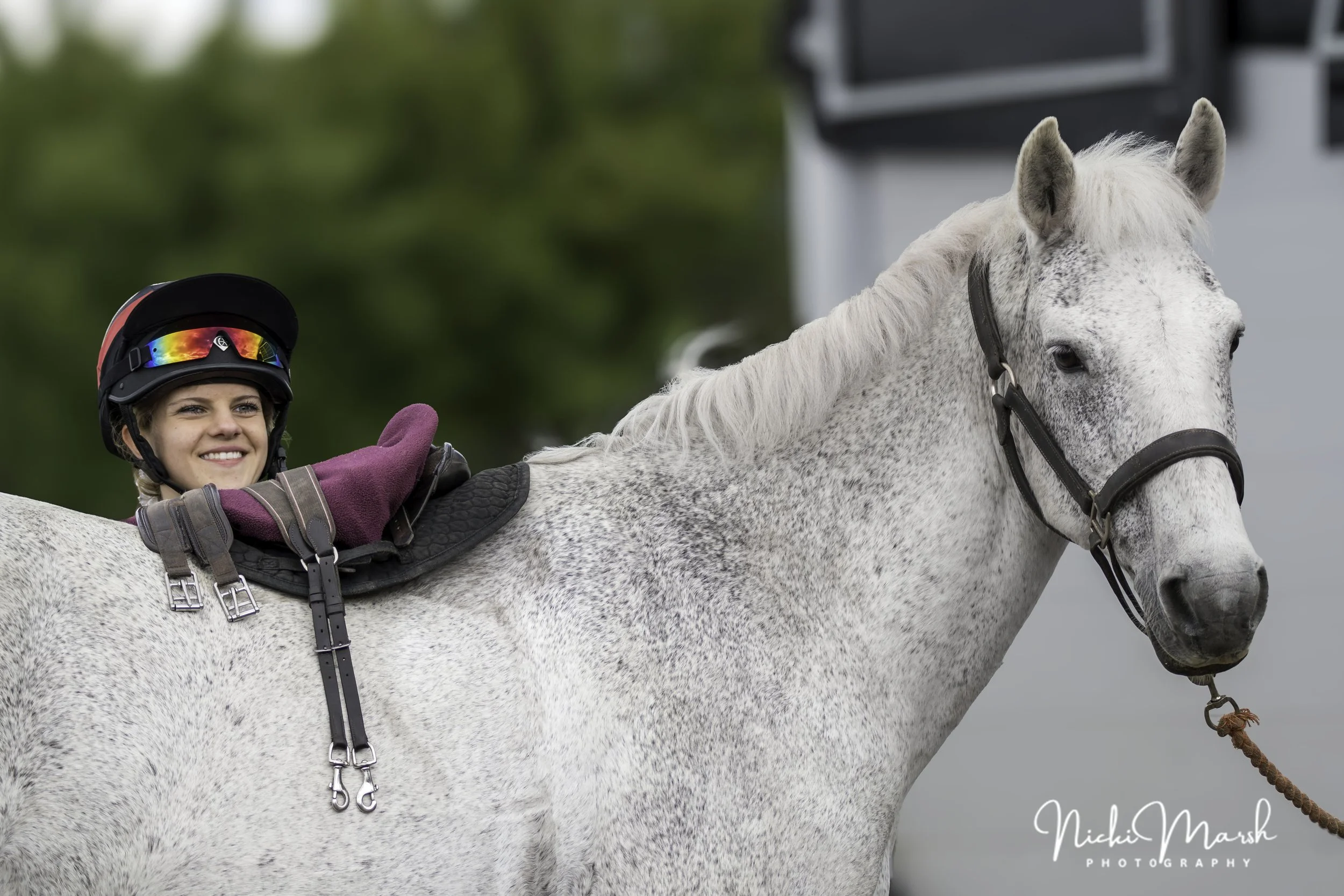 A smiling woman wearing a black riding helmet with rainbow-colored reflective sunglasses on top, pink gloves, and a riding jacket, riding a white horse with gray speckles on its neck and face. The horse is wearing a black bridle and saddle. The backg