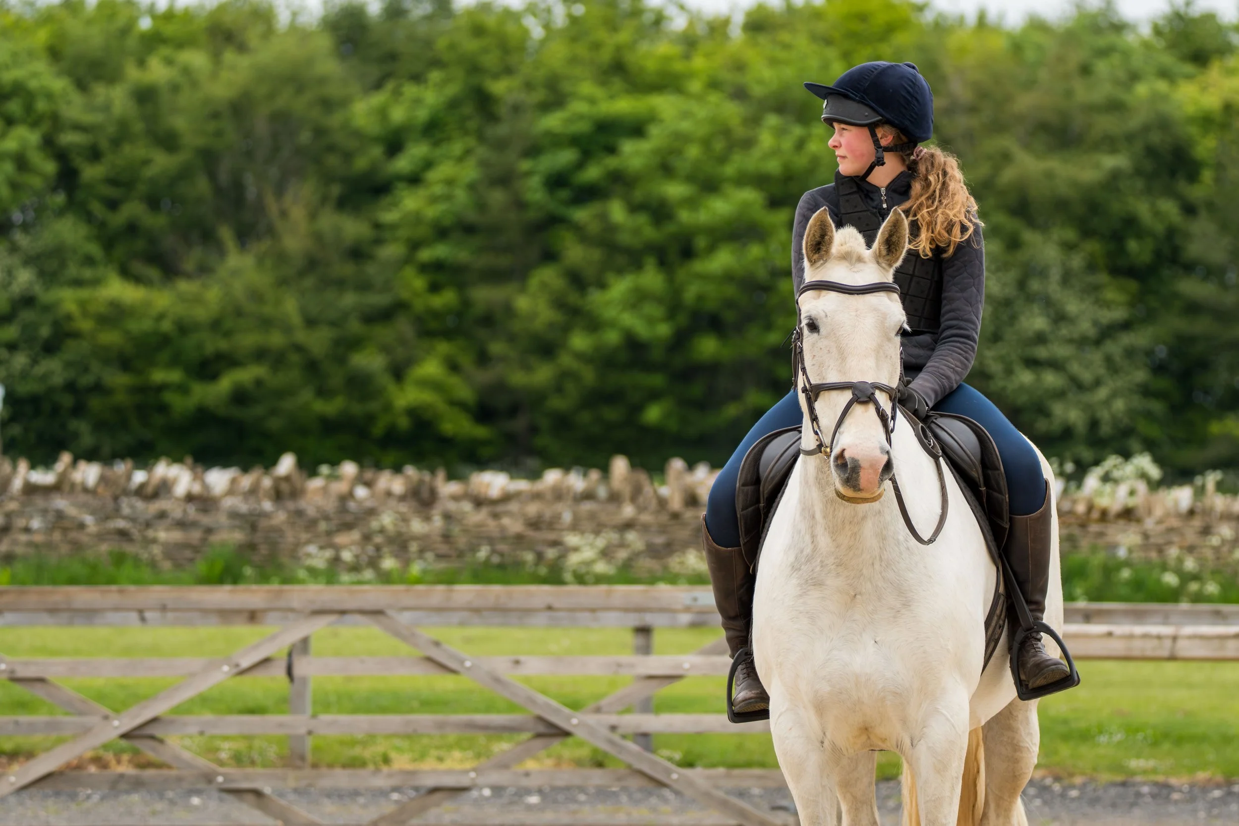 A young woman with curly hair wearing a helmet and riding gear, riding a white horse in an outdoor riding arena with a wooden fence and green trees in the background.