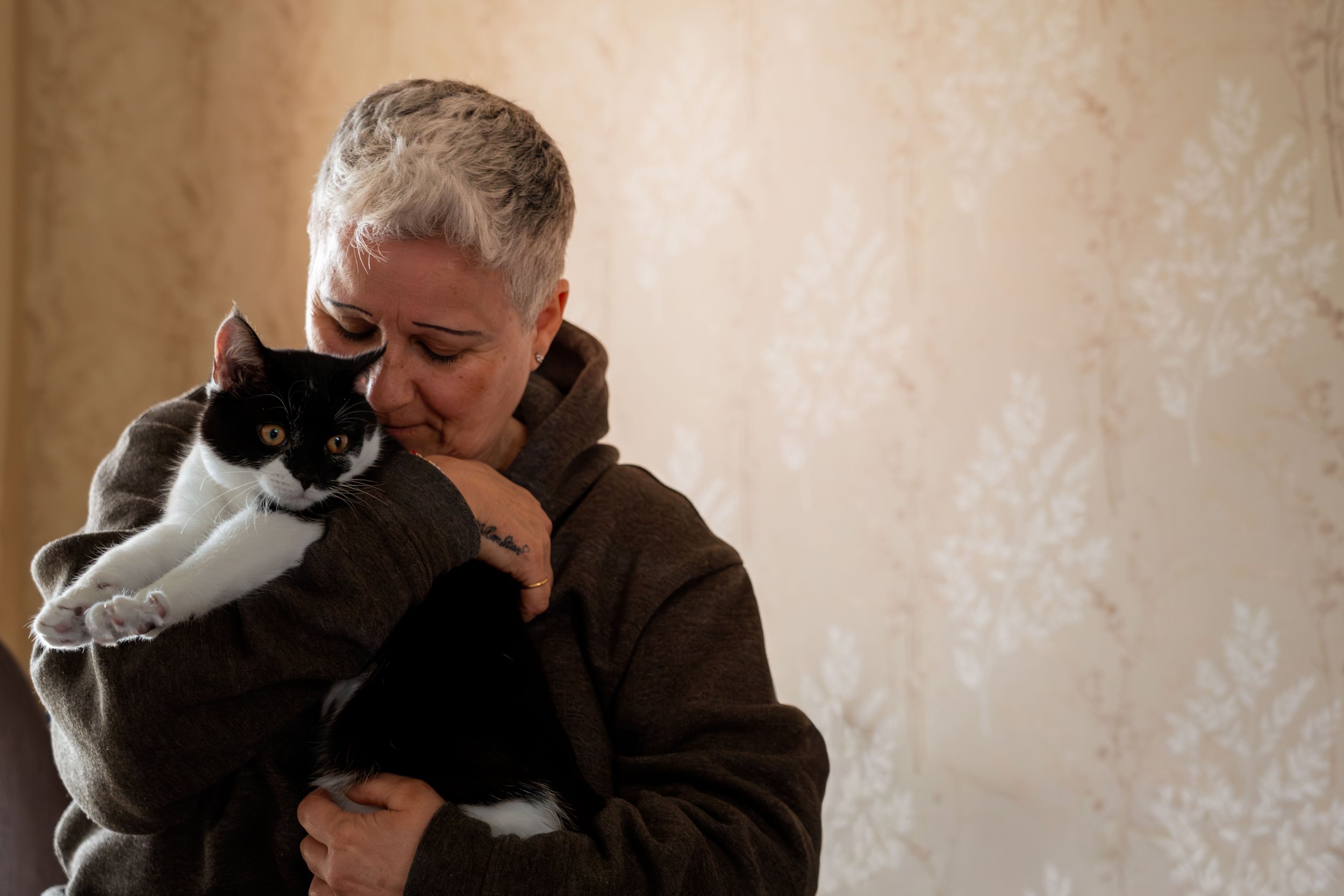 A person with short blonde hair holding a black and white cat close to their face, both with eyes open, in front of a beige textured wall.