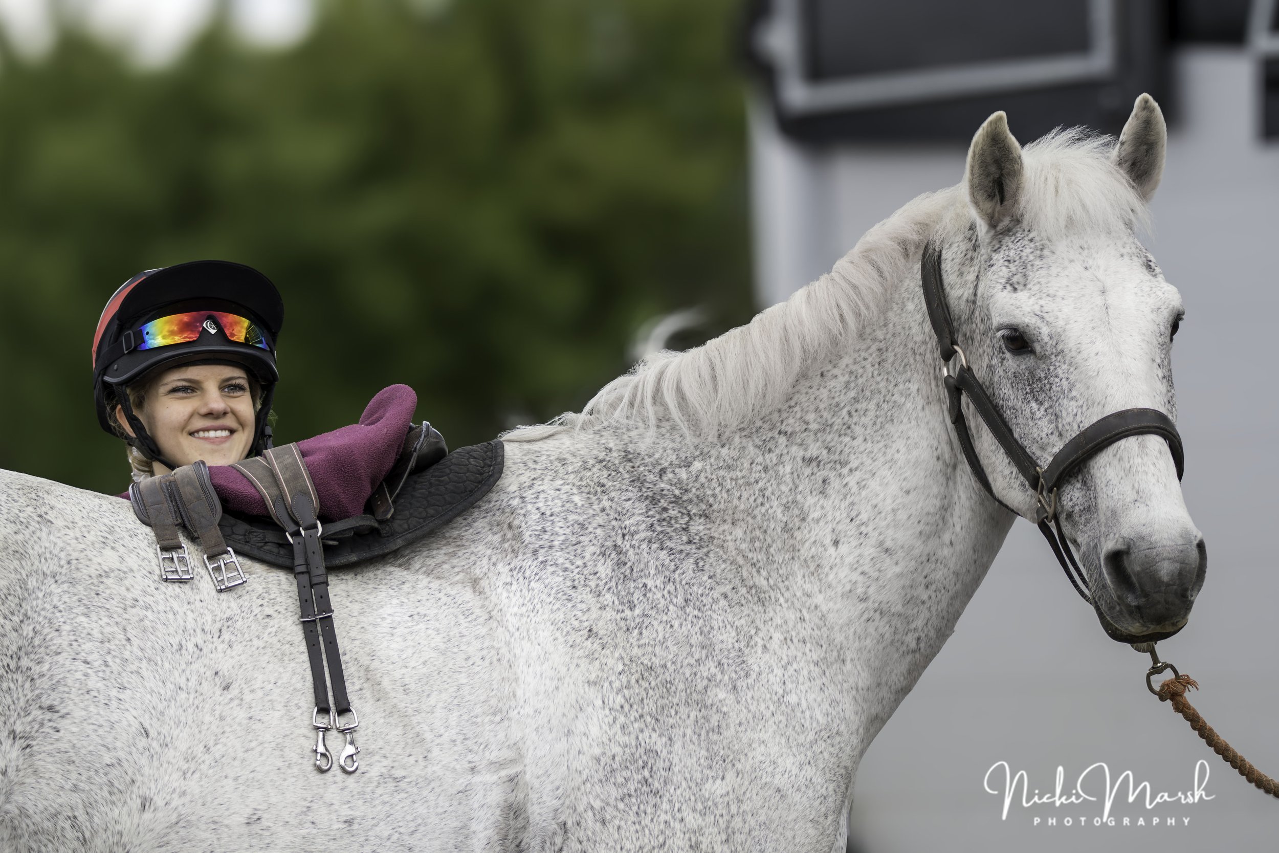 A young woman with a helmet and gloves riding a white horse with gray spots outdoors, smiling and giving a thumbs-up.