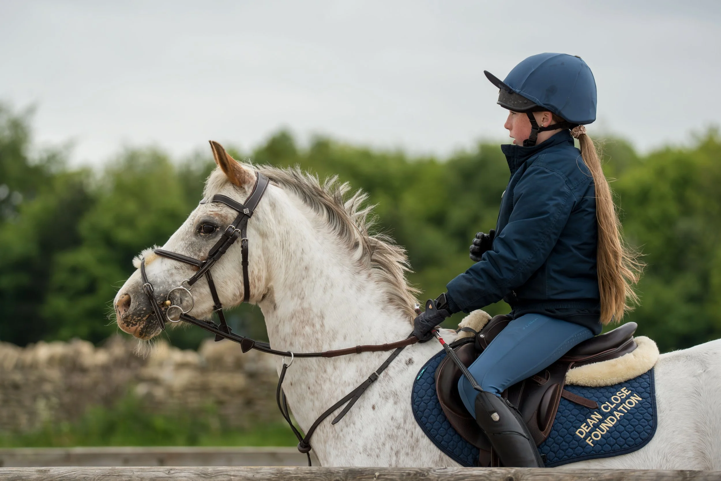 A young woman with a long ponytail riding a white horse in an outdoor setting with green trees and a cloudy sky.