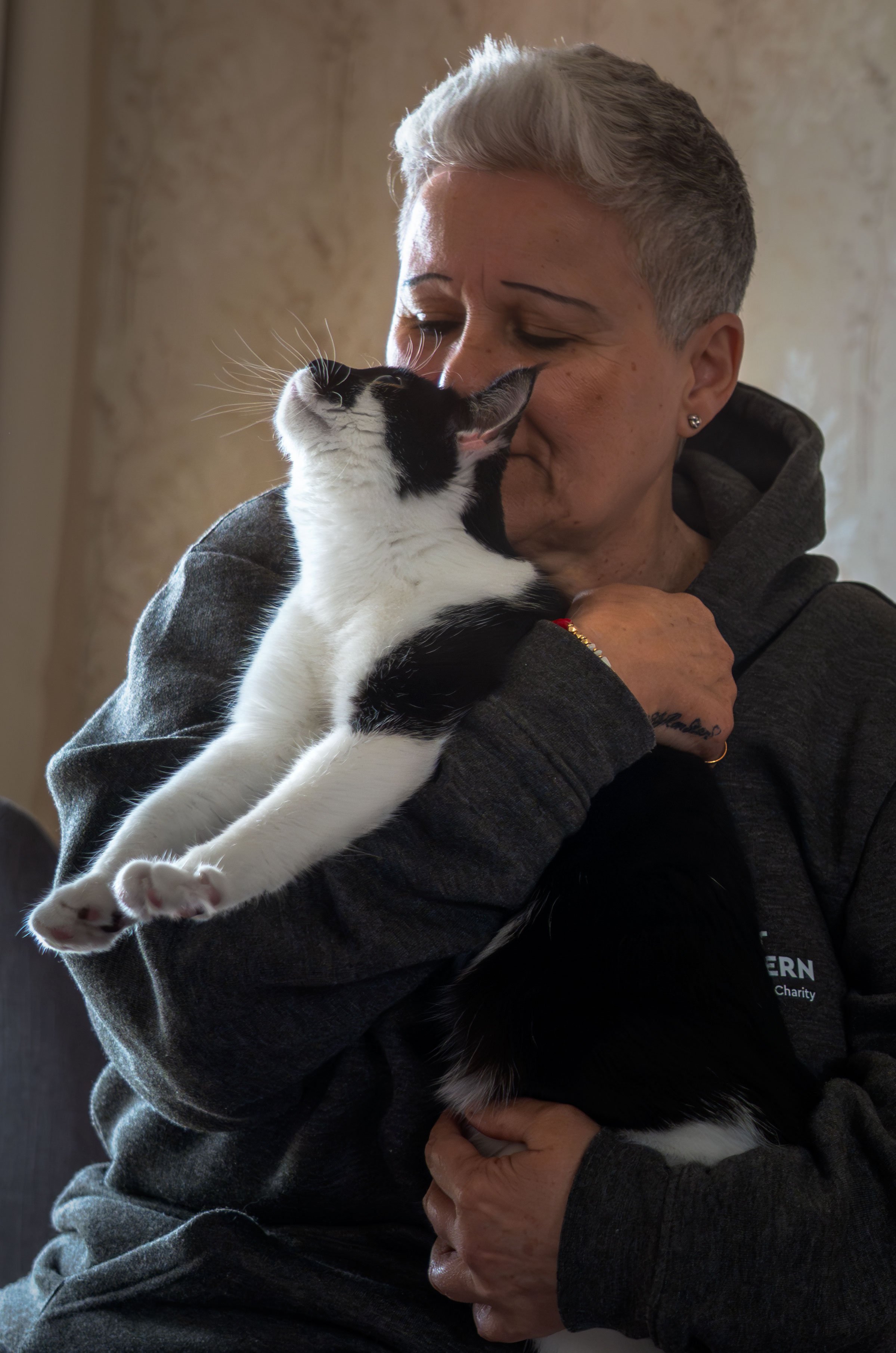 A person with short gray hair and earrings holding a black and white cat close to their face, the cat appears to be sniffing or nuzzling the person's nose. The person is wearing a dark hoodie and the background is neutral.