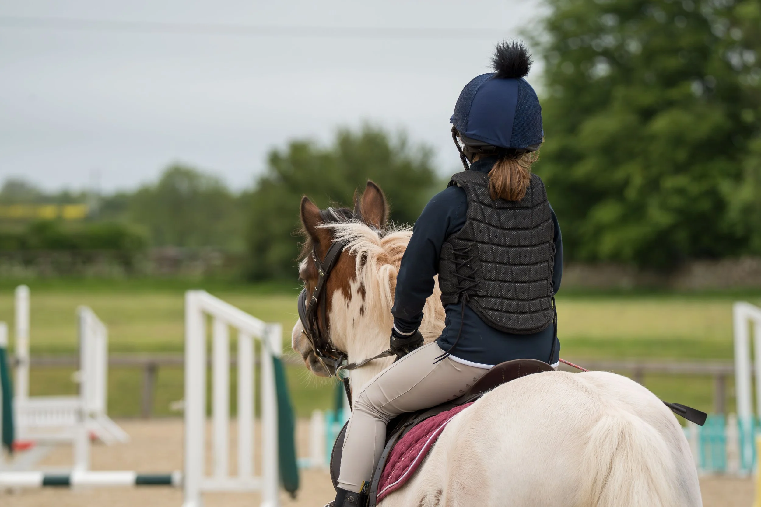 A person riding a white horse during a trail ride, wearing a helmet and a black vest, with a green outdoor background.