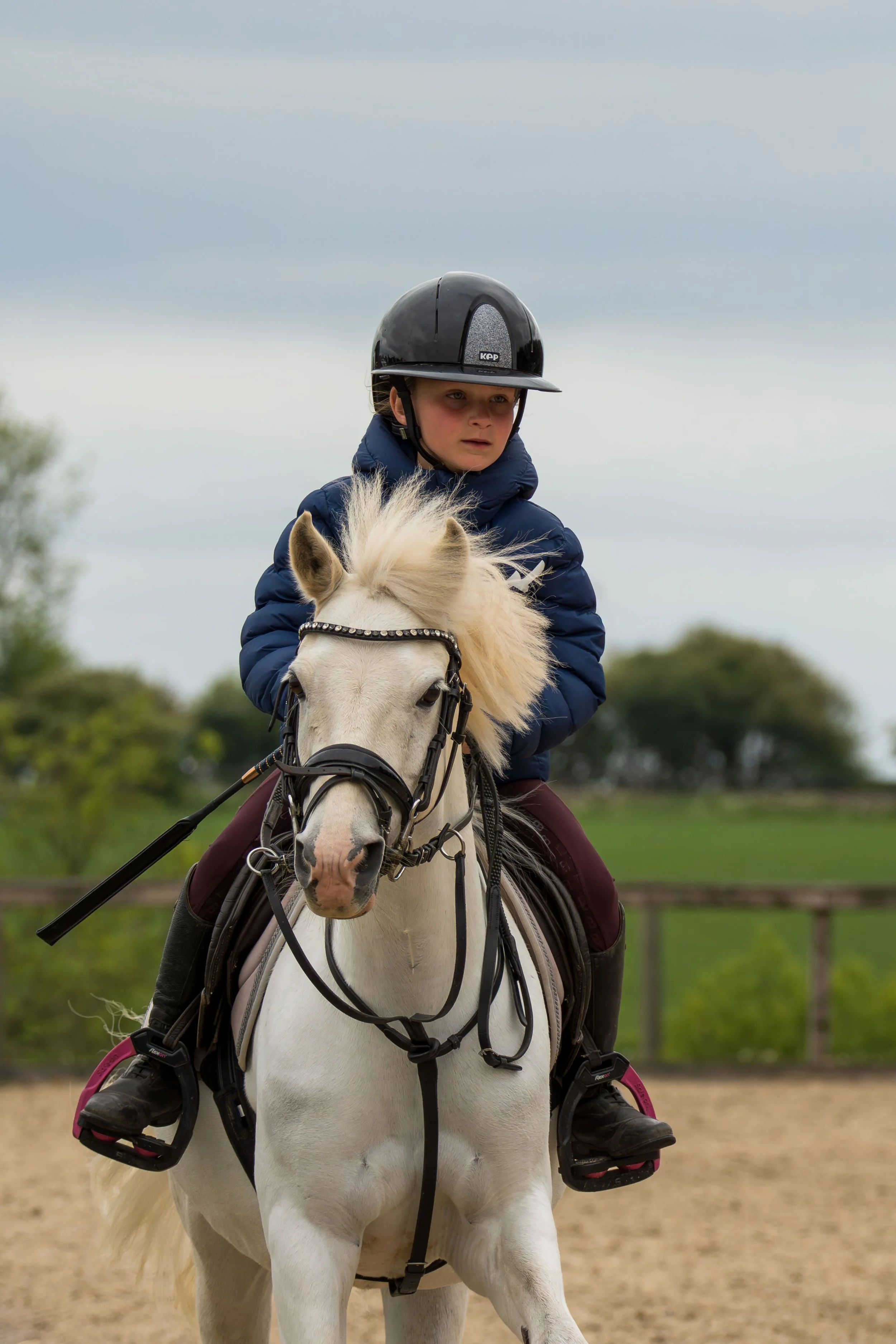 A young girl in a helmet riding a white horse in an outdoor riding arena with a cloudy sky.