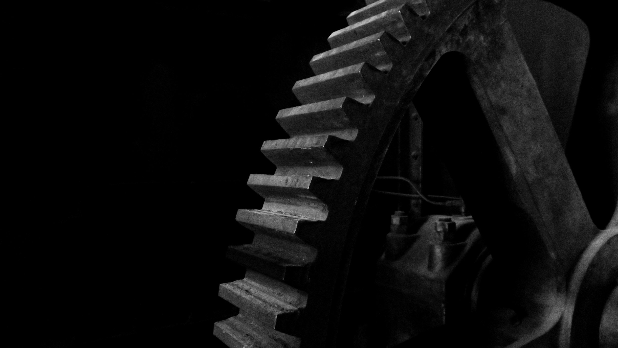 A close-up view of a large industrial gear with blocky teeth, mounted on a dark background, in black and white.