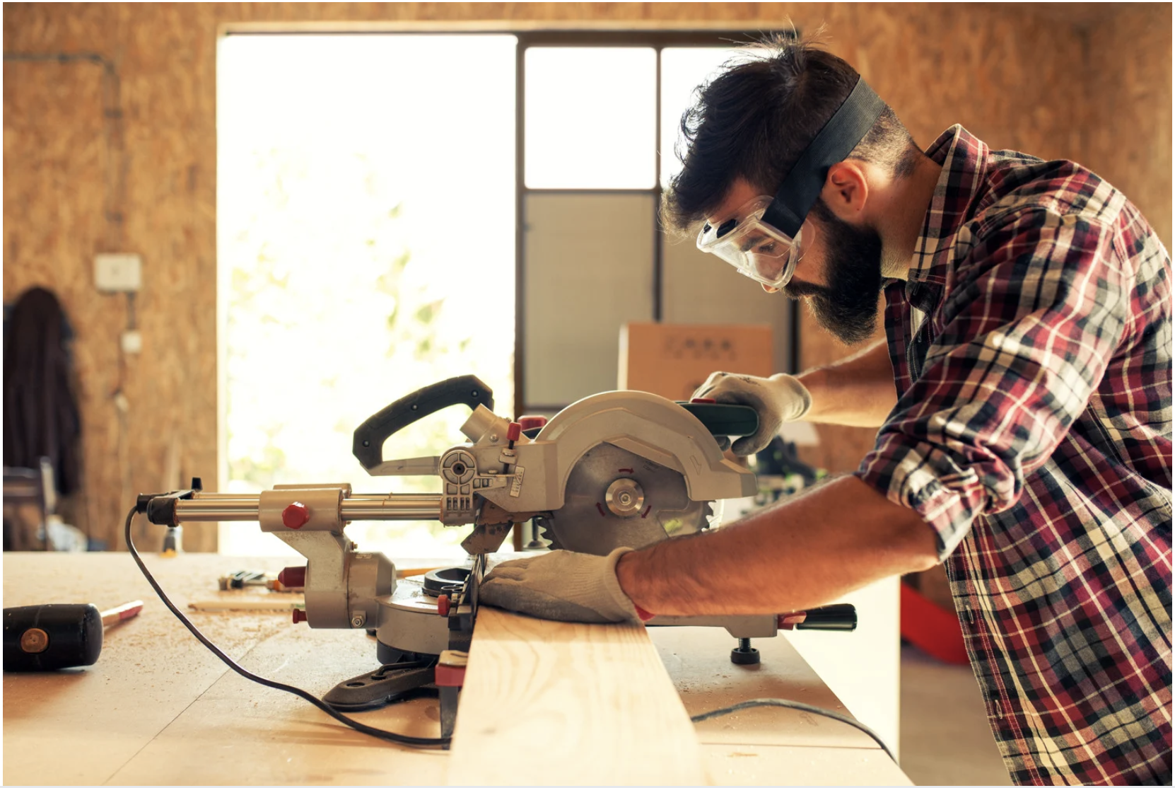 A man wearing safety goggles, a plaid shirt, and gloves uses a circular saw to cut a piece of wood in a woodworking workshop with a brick wall background.