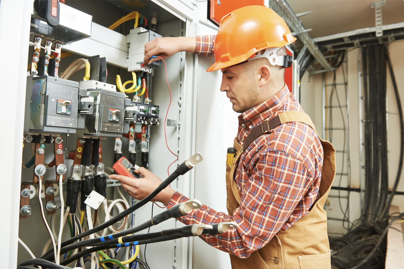 An electrician wearing an orange hard hat and plaid shirt inspecting an electrical panel with a multimeter in hand in an industrial setting.