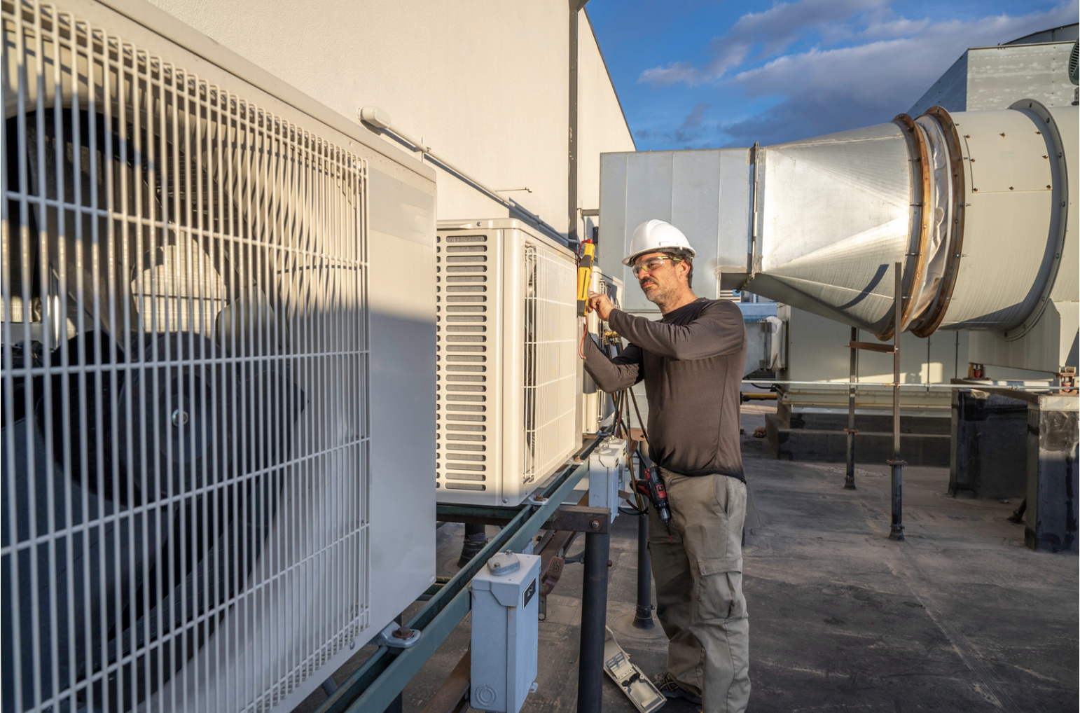 A technician wearing a white safety helmet and glasses working on an HVAC unit on a roof, using a multimeter connected to the equipment.