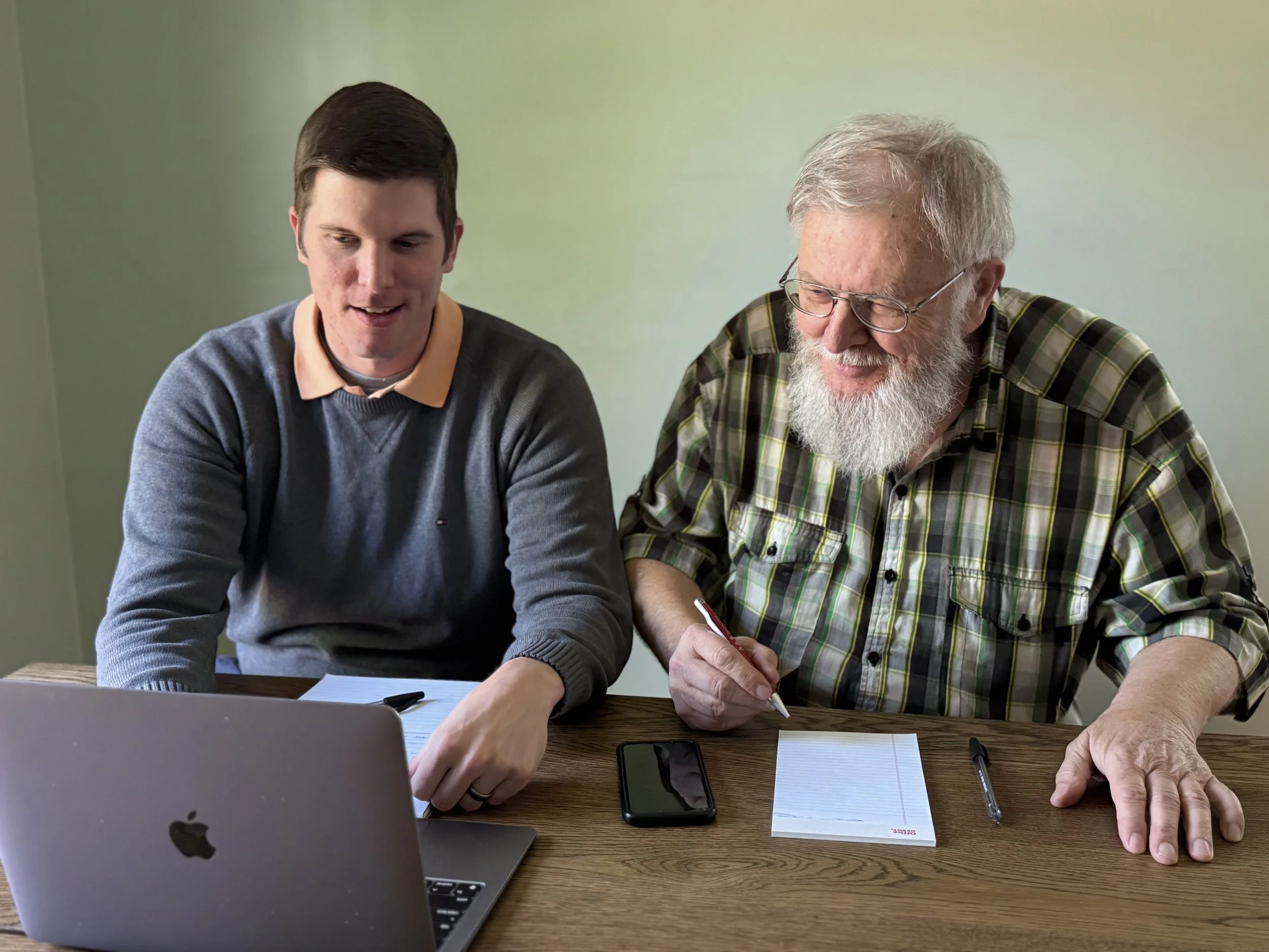 Two men, one young and one older with glasses and a beard, sit at a wooden table with papers, pen, and smartphones, engaging in discussion in a room with green walls and a closed laptop in the foreground.