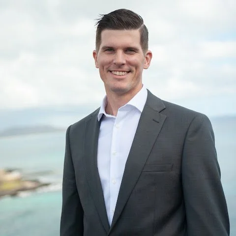 A smiling man in a gray suit with a white shirt stands outdoors with a body of water and cloudy sky in the background.