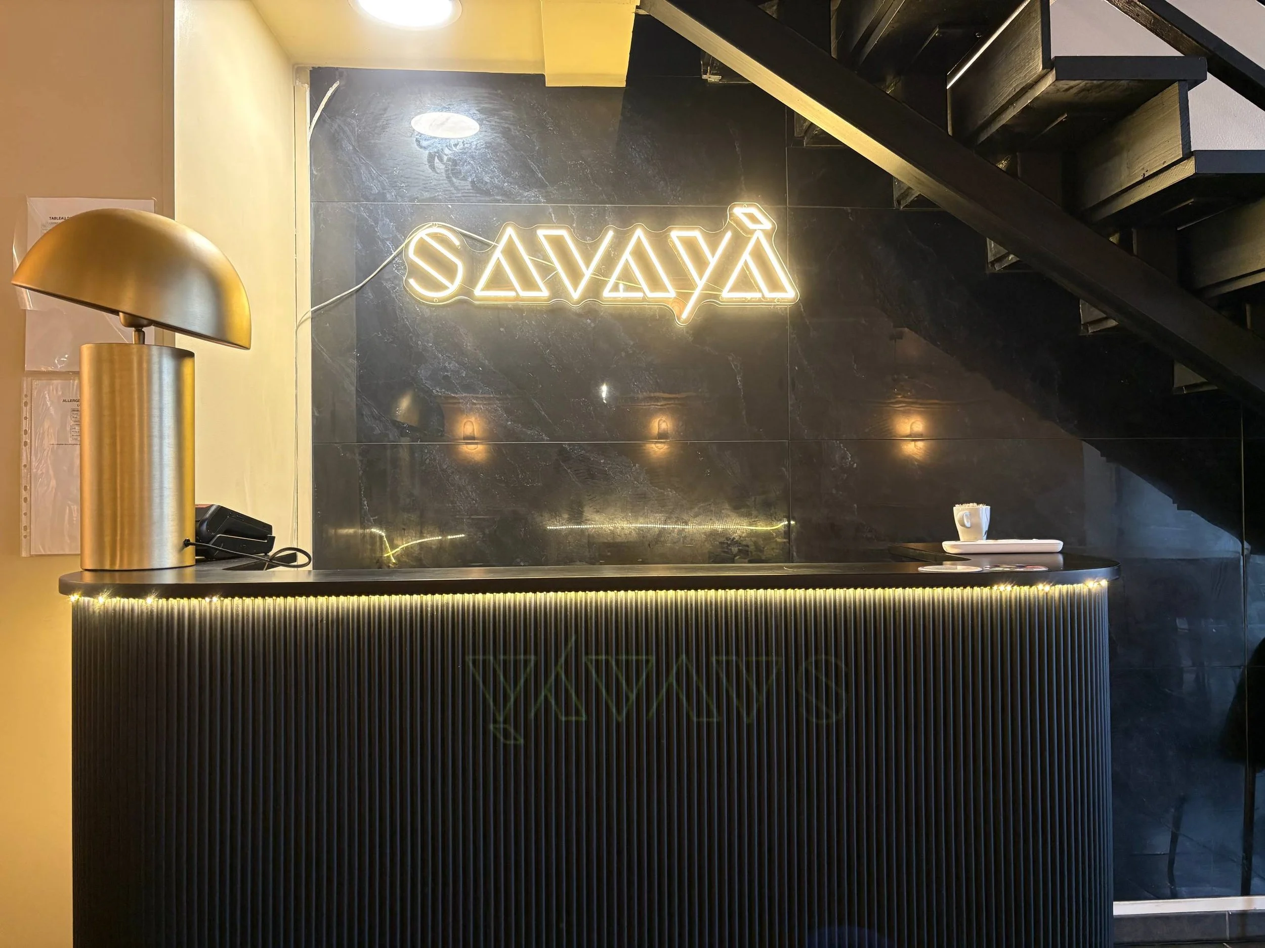 Une enseigne lumineuse indiquant 'SAVAYA' accrochée sur un mur noir brillant, avec un éclairage chaud et une table avec une tasse en porcelaine blanche et des petits objets. Un lampadaire métallique doré est aussi visible à gauche.
