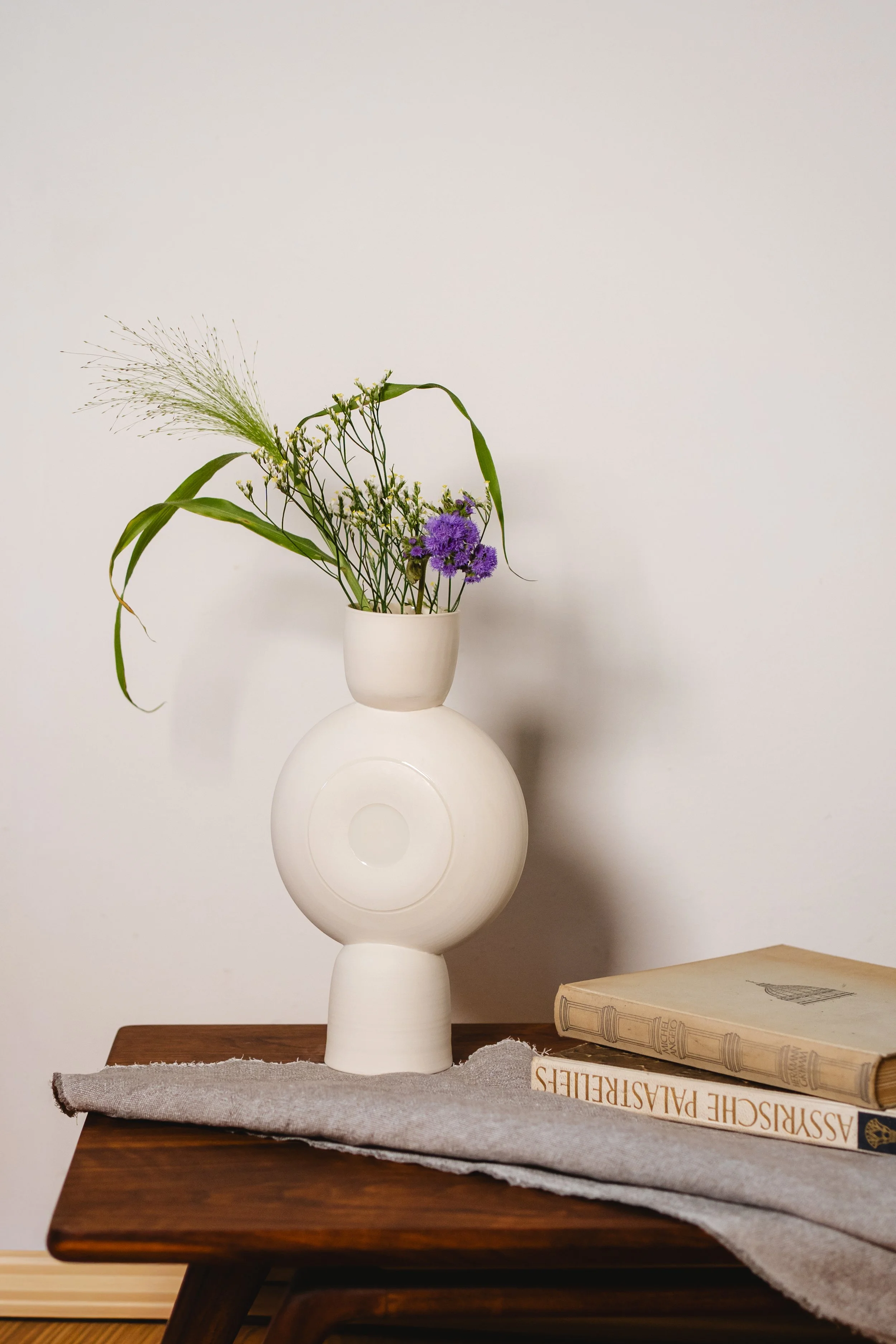 weisse skulpturale Vase steht auf Holztisch mit zwei Büchern und Tischdecke.

