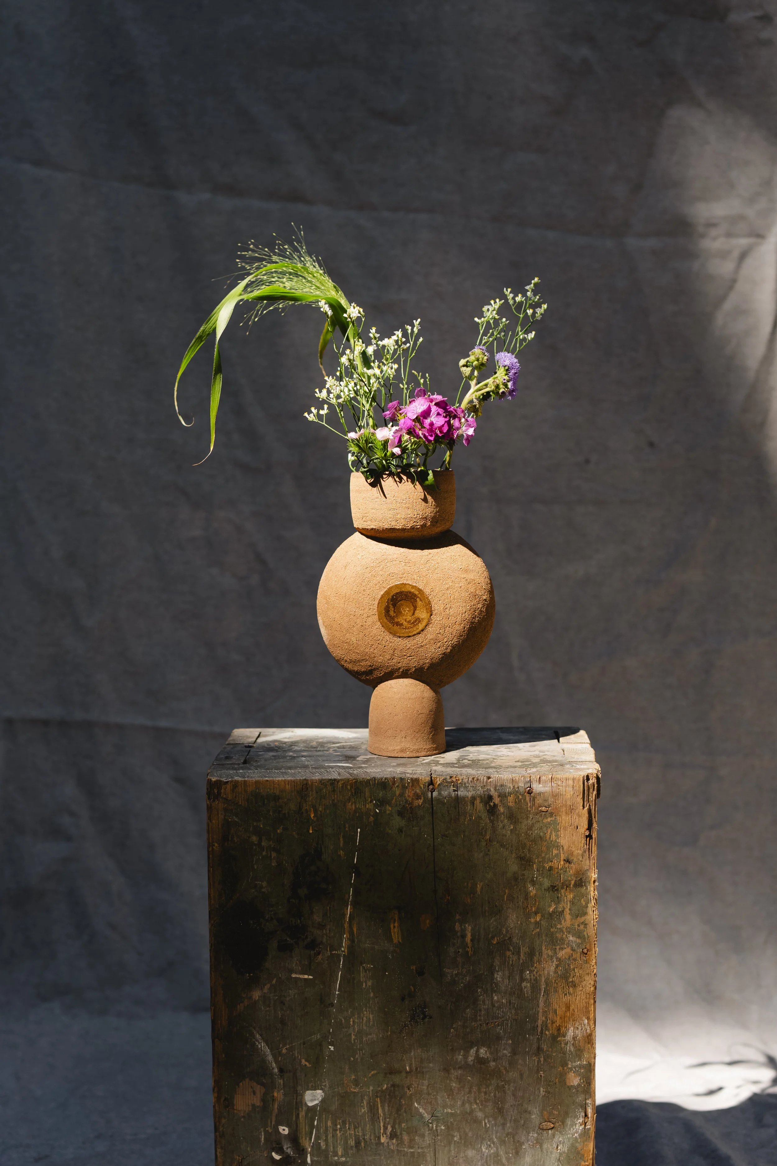 Skulpturale braune Vase mit Blumen steht auf einem Holzpodest vor grauem Hintergrund.