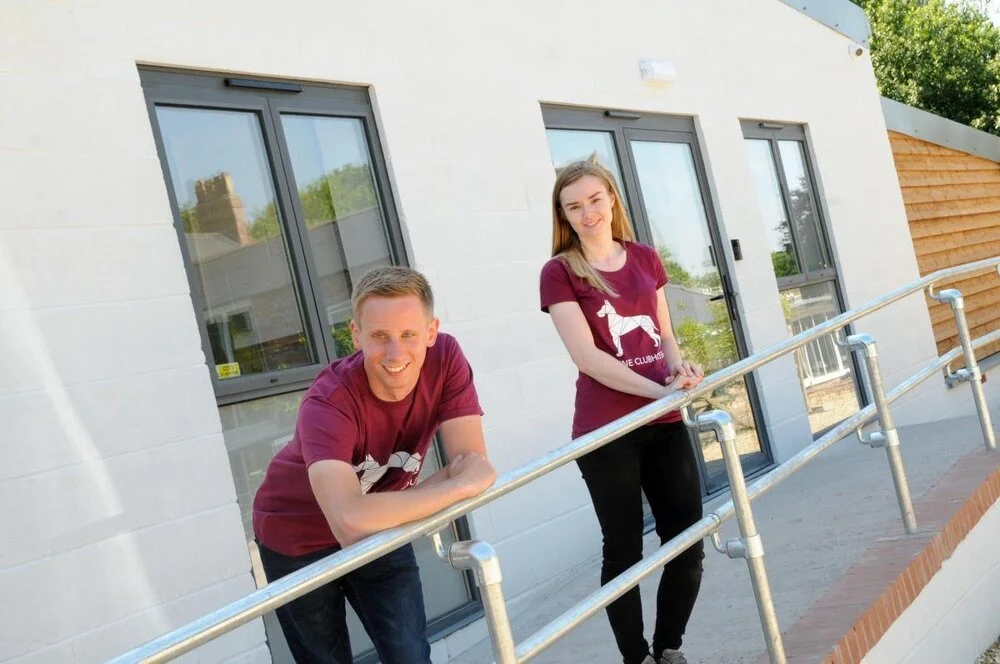 Two young adults, a man and a woman, leaning on a metal railing outside a modern building with large windows. They are smiling and wearing matching maroon t-shirts with a white alligator graphic.