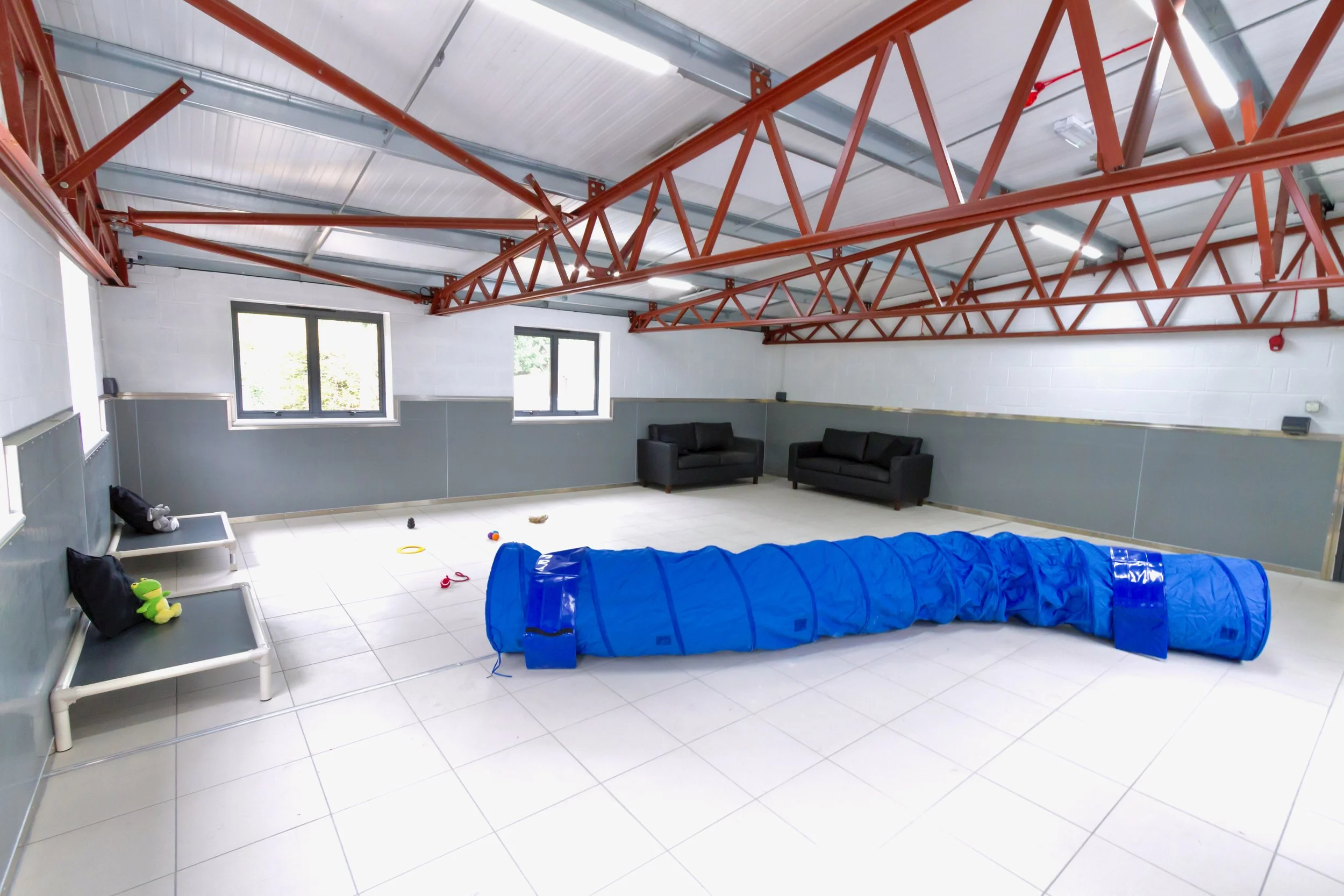 An indoor playroom with two black sofas, a blue play tunnel, and various toys scattered on a tiled floor. There are two windows on the wall, metal and red beams on the ceiling, and white walls with gray paneling.