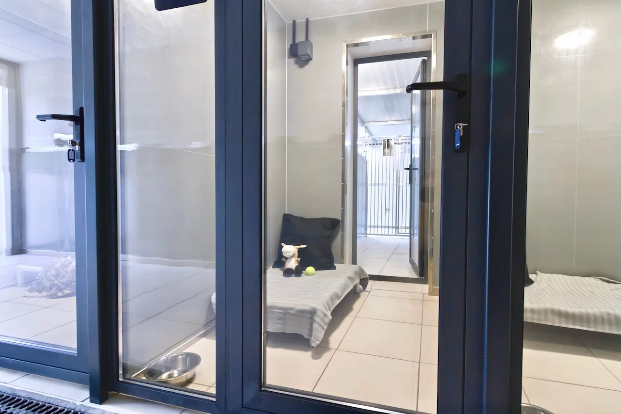 A small indoor animal enclosure with a glass door, a small bed with a stuffed animal, a tennis ball, and a metal food bowl, with a balcony in the background.