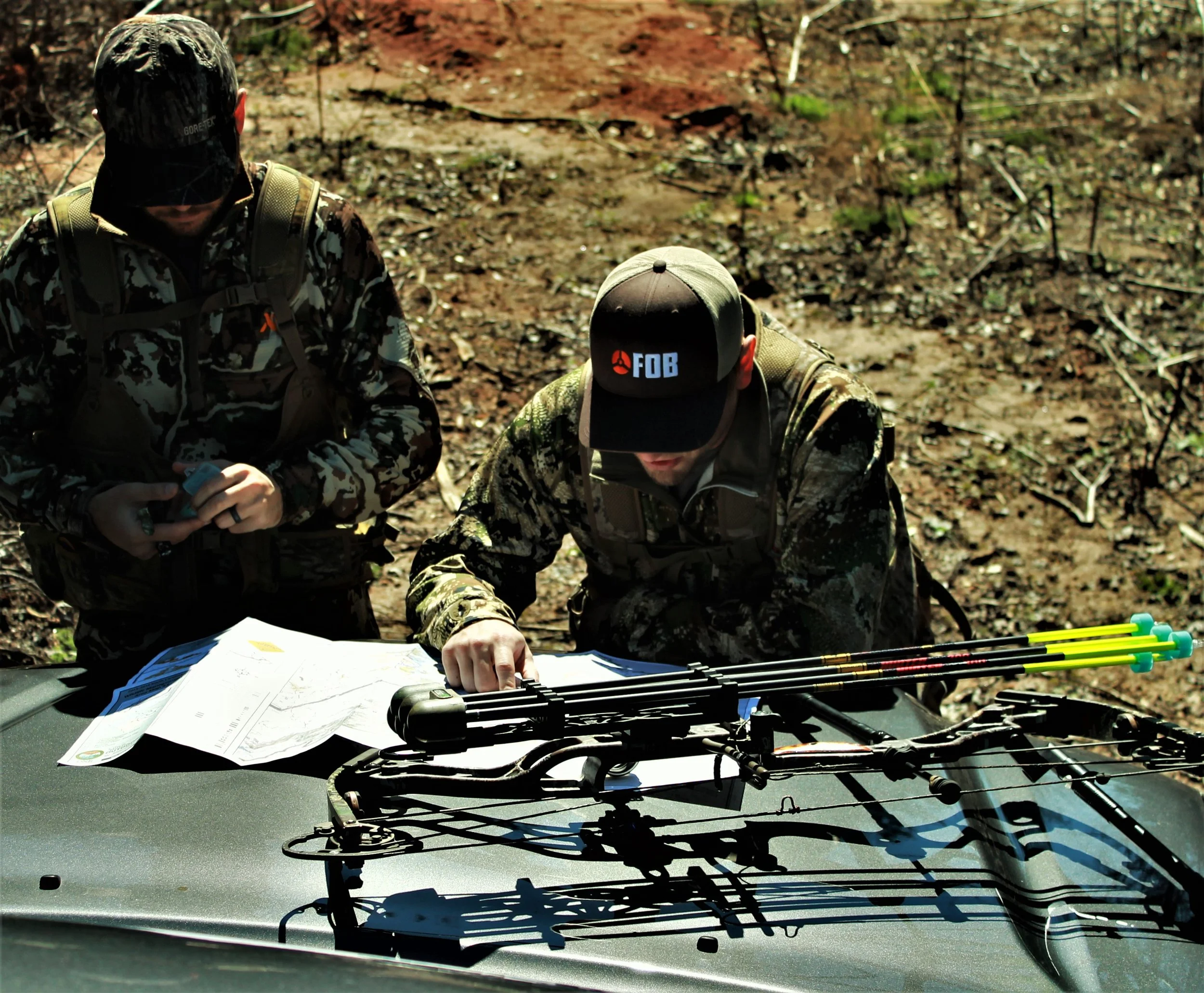 Two men in camouflage clothing and caps sitting outdoors at a table with archery equipment, maps, and documents.